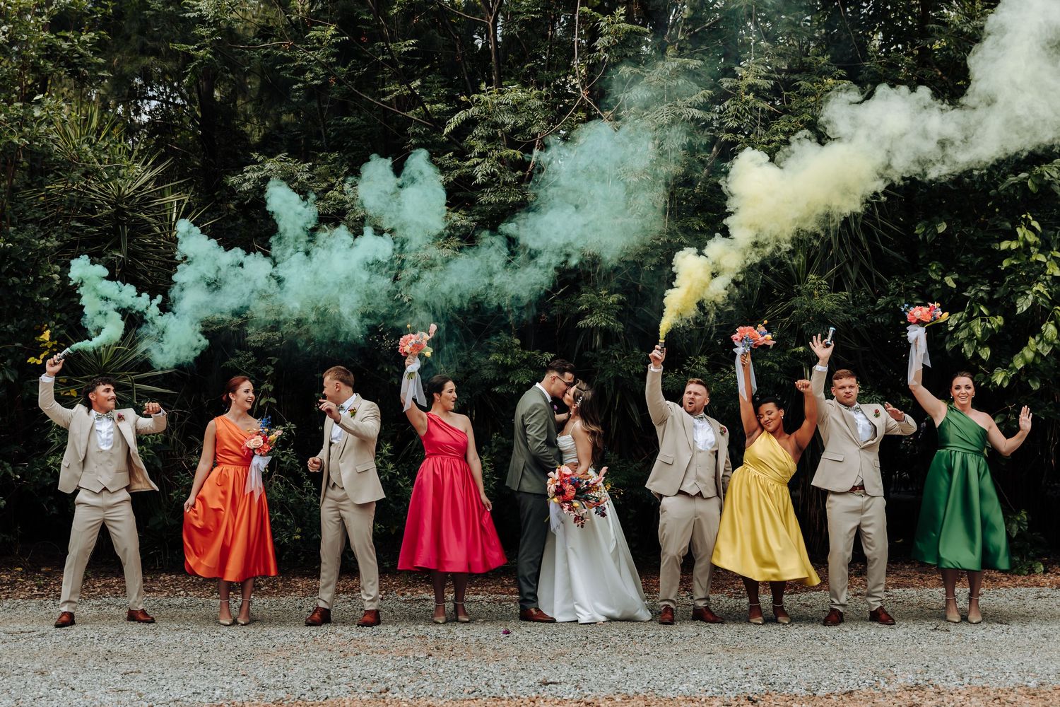Wedding party poses with colorful smoke bombs creating a rainbow effect against a wooded backdrop.