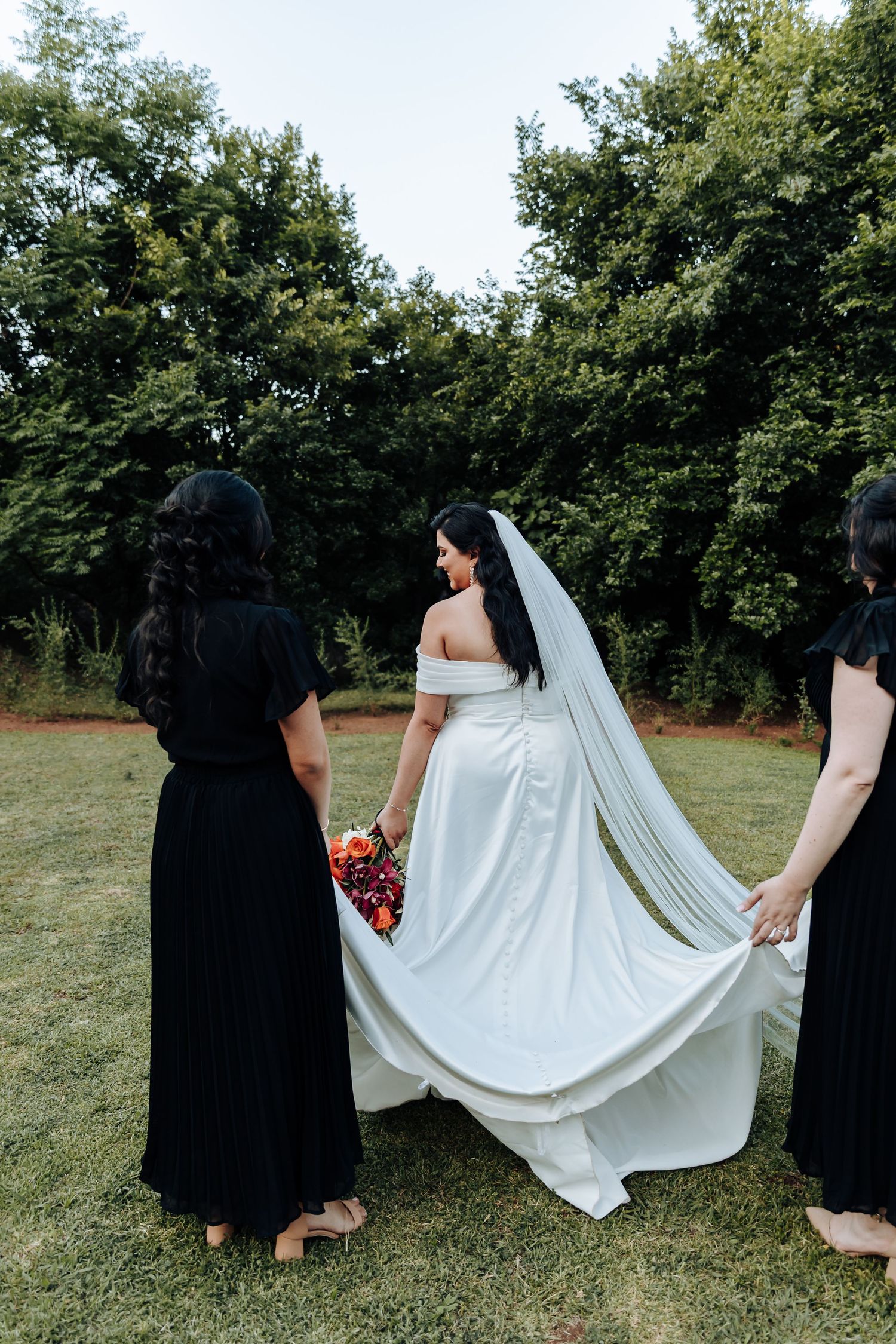 Bridesmaids in black dresses help arrange the bride's flowing white dress train on grass.