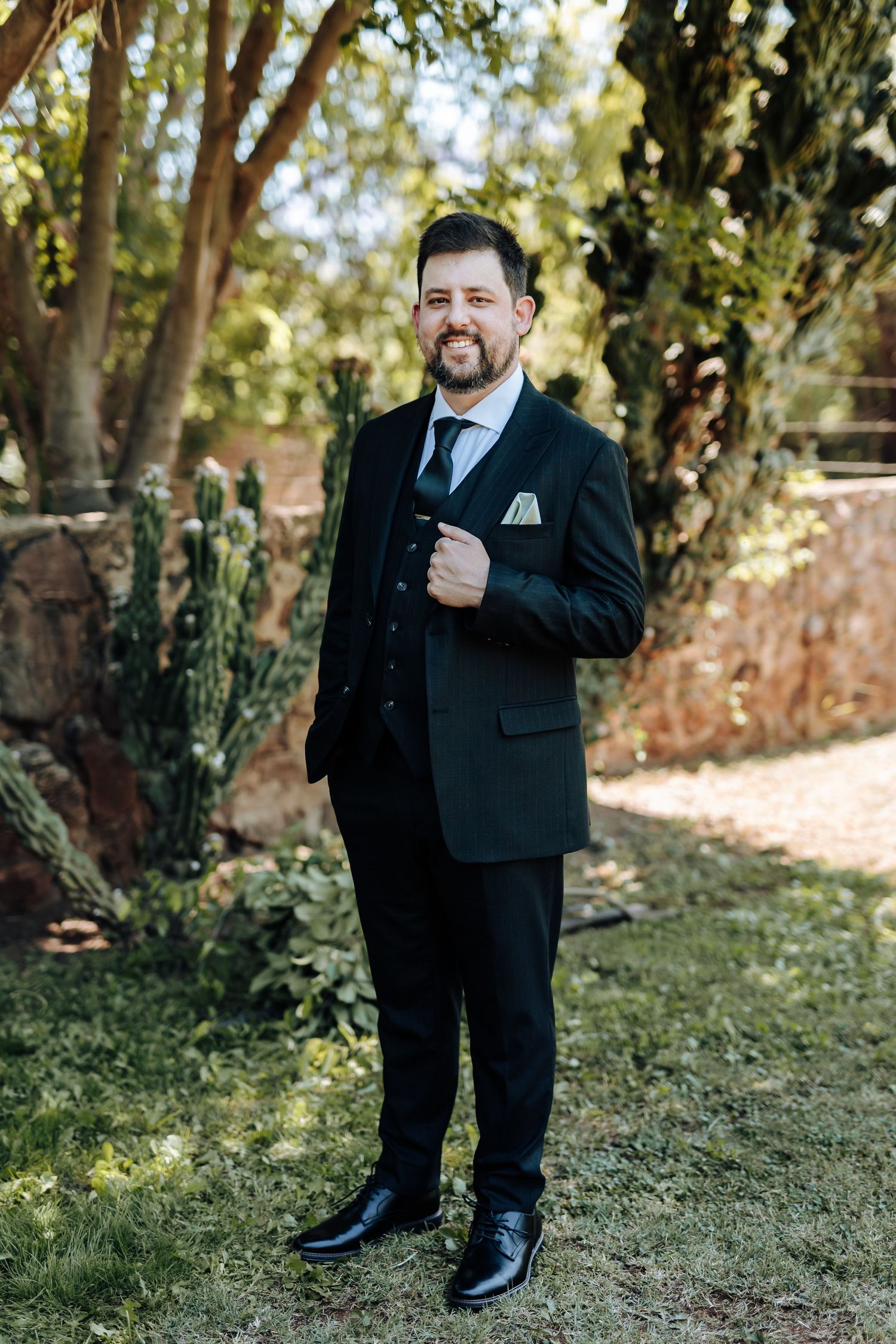Person in black suit and tie posing outdoors in front of trees and brick wall.