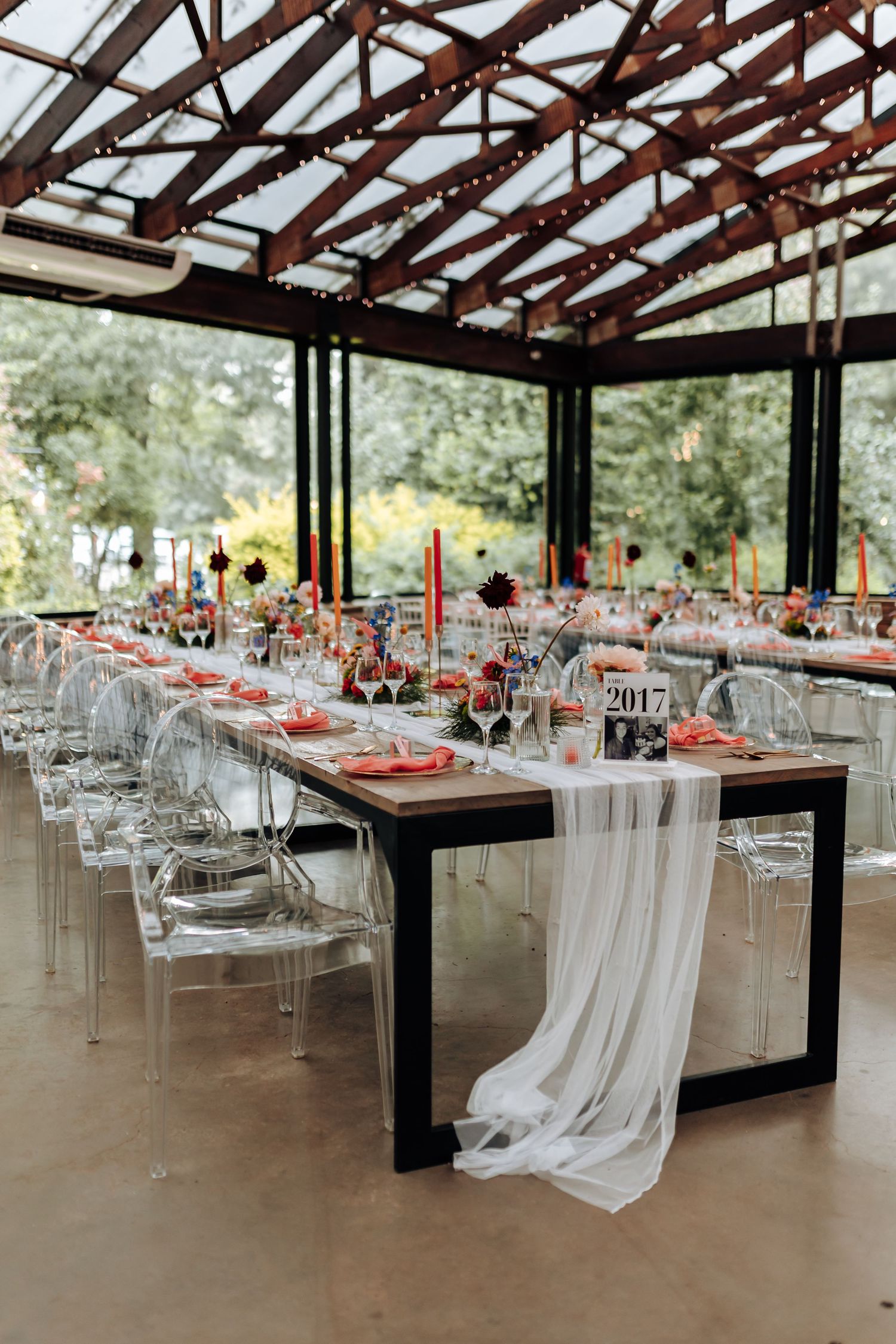 Modern reception venue with clear ghost chairs and long tables set with red accents under a rustic wooden pergola ceiling.