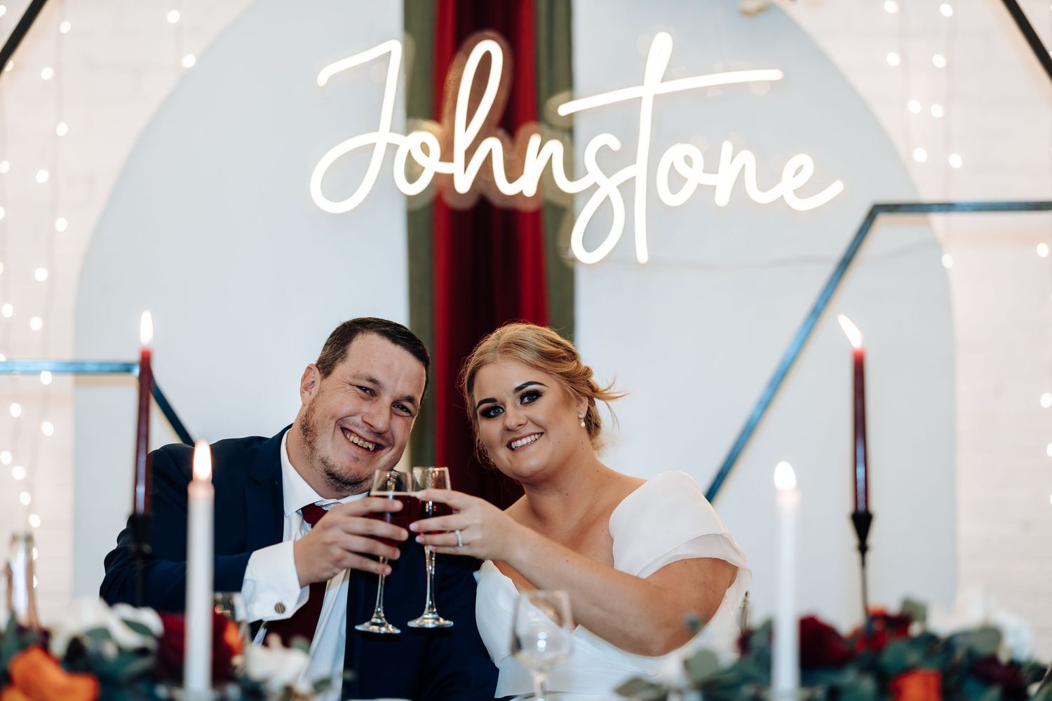 Newlywed couple toasting with wine glasses in front of a glowing Johnstone neon sign at their wedding reception.