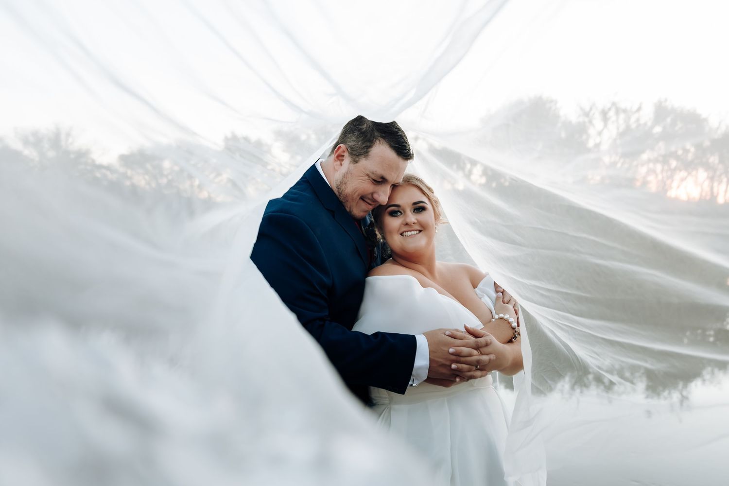 Bride and groom embrace tenderly under a flowing white veil on their wedding day.