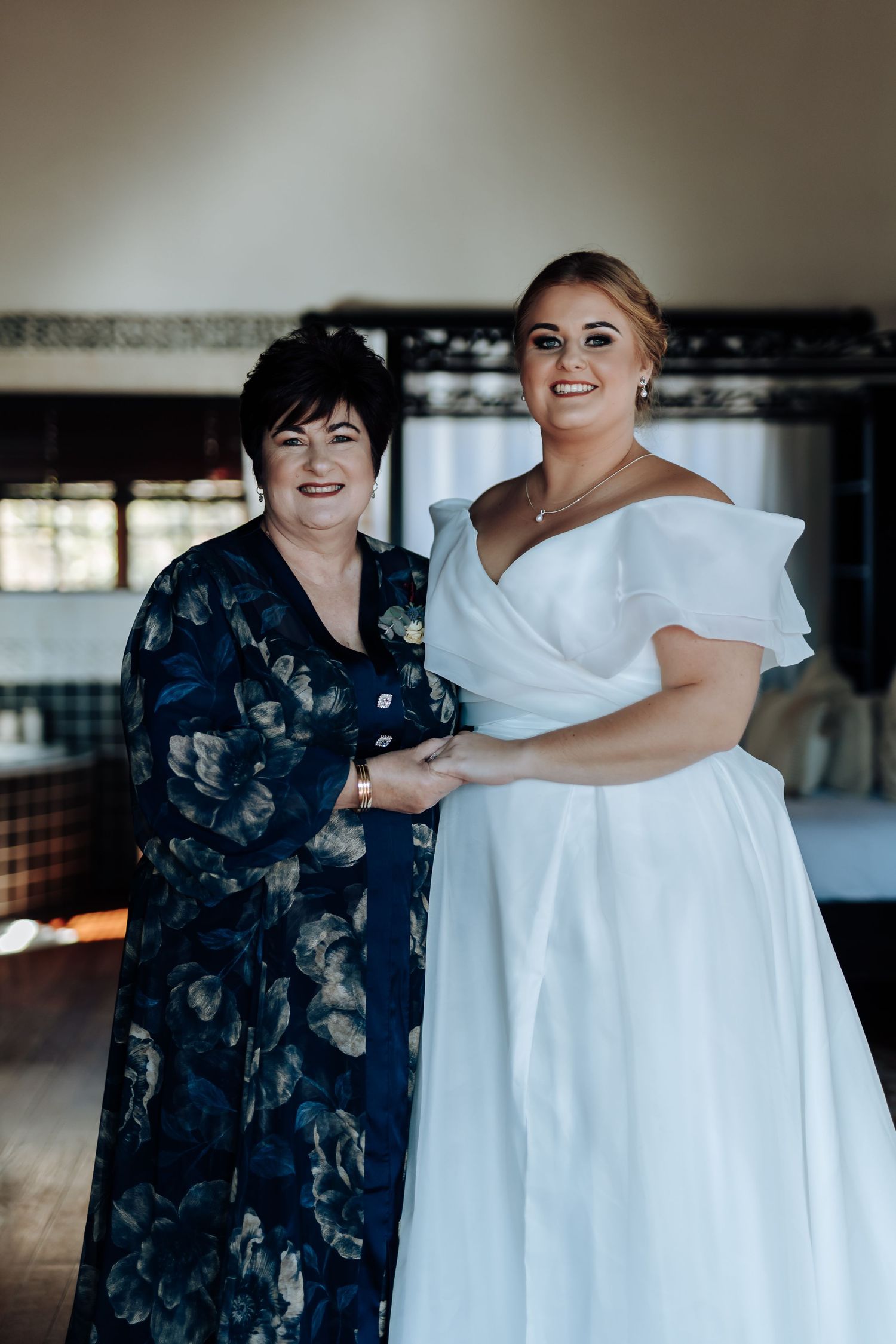 A bride in a white off-shoulder gown poses with an older woman in a blue floral dress, holding hands and smiling.