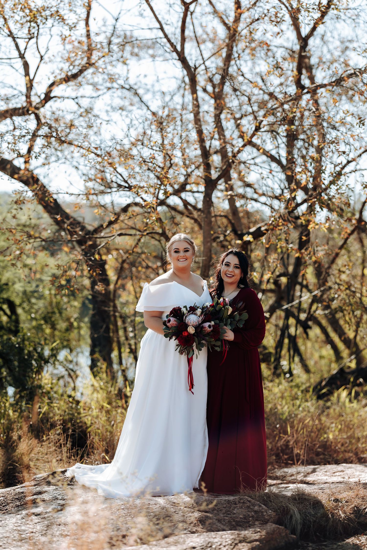 Bride in white gown and bridesmaid in burgundy dress holding bouquets outdoors among autumn trees.