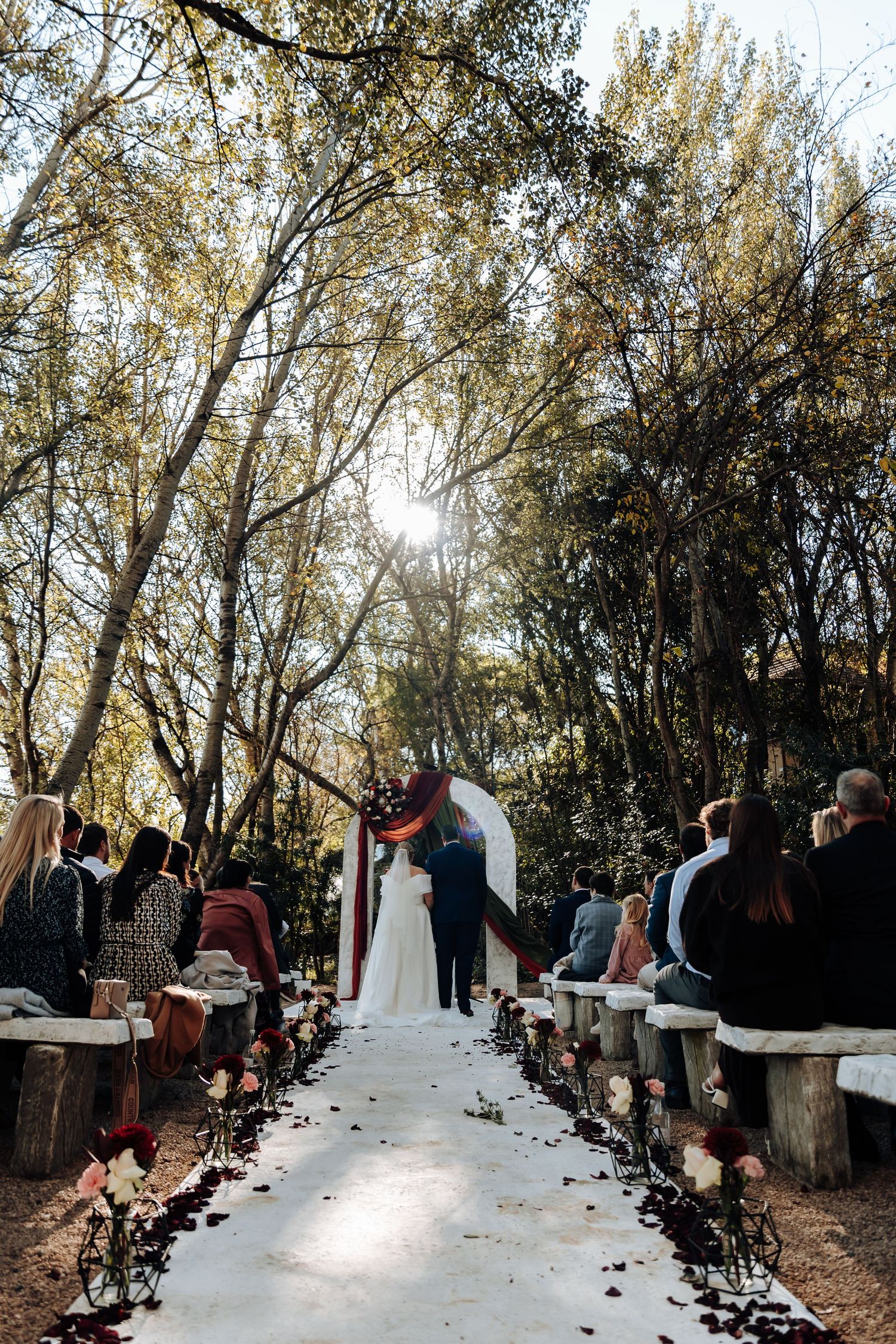 Outdoor wedding ceremony aisle lined with rose petals and rustic benches under sunlit trees with couple at floral arch.
