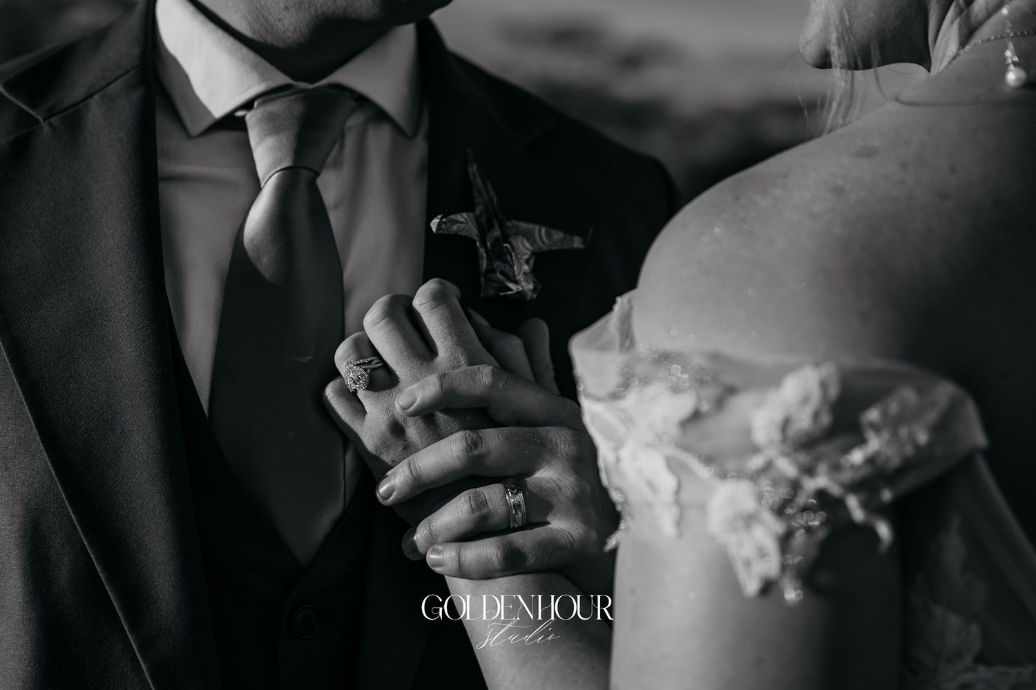 Black and white photo of bride and groom holding hands, showcasing wedding rings and boutonniere.
