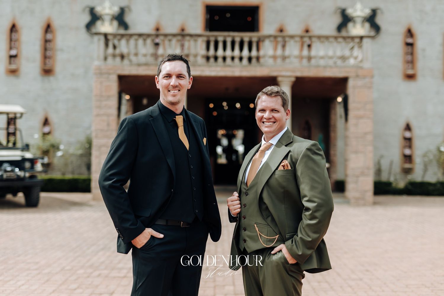 Two grooms in elegant suits pose together outside a castle venue on their wedding day.