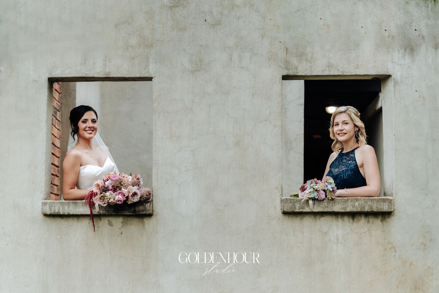 Bride and bridesmaid in navy dress each posing in separate stone wall windows holding bouquets.