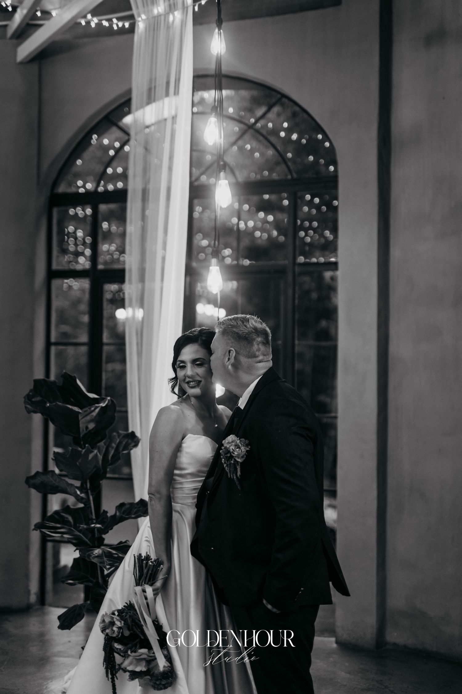 Black and white portrait of bride and groom sharing a tender moment under draped fabric and fairy lights.