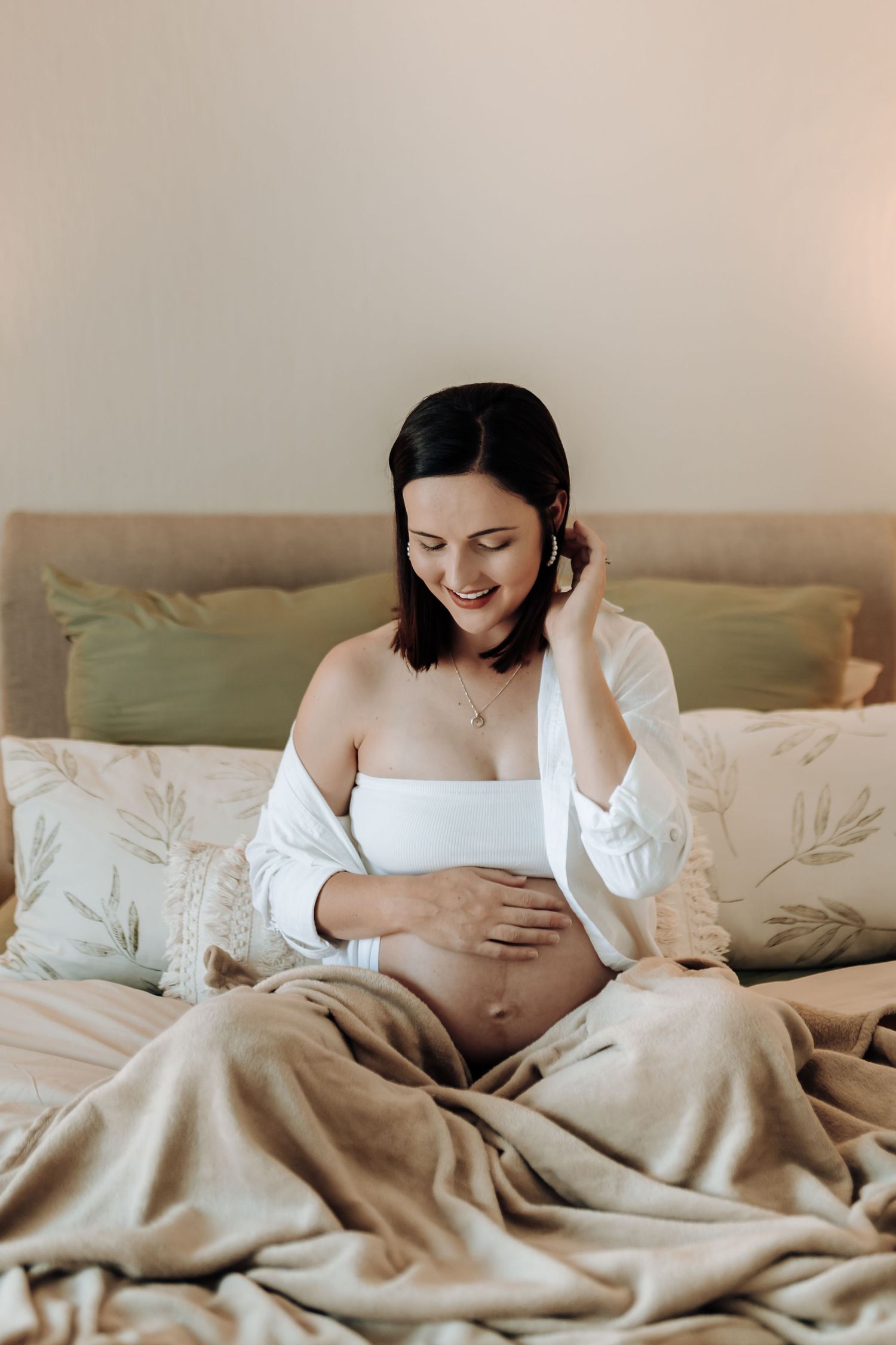 Pregnant woman in white shirt reclines on bed with blanket in soft natural lighting.