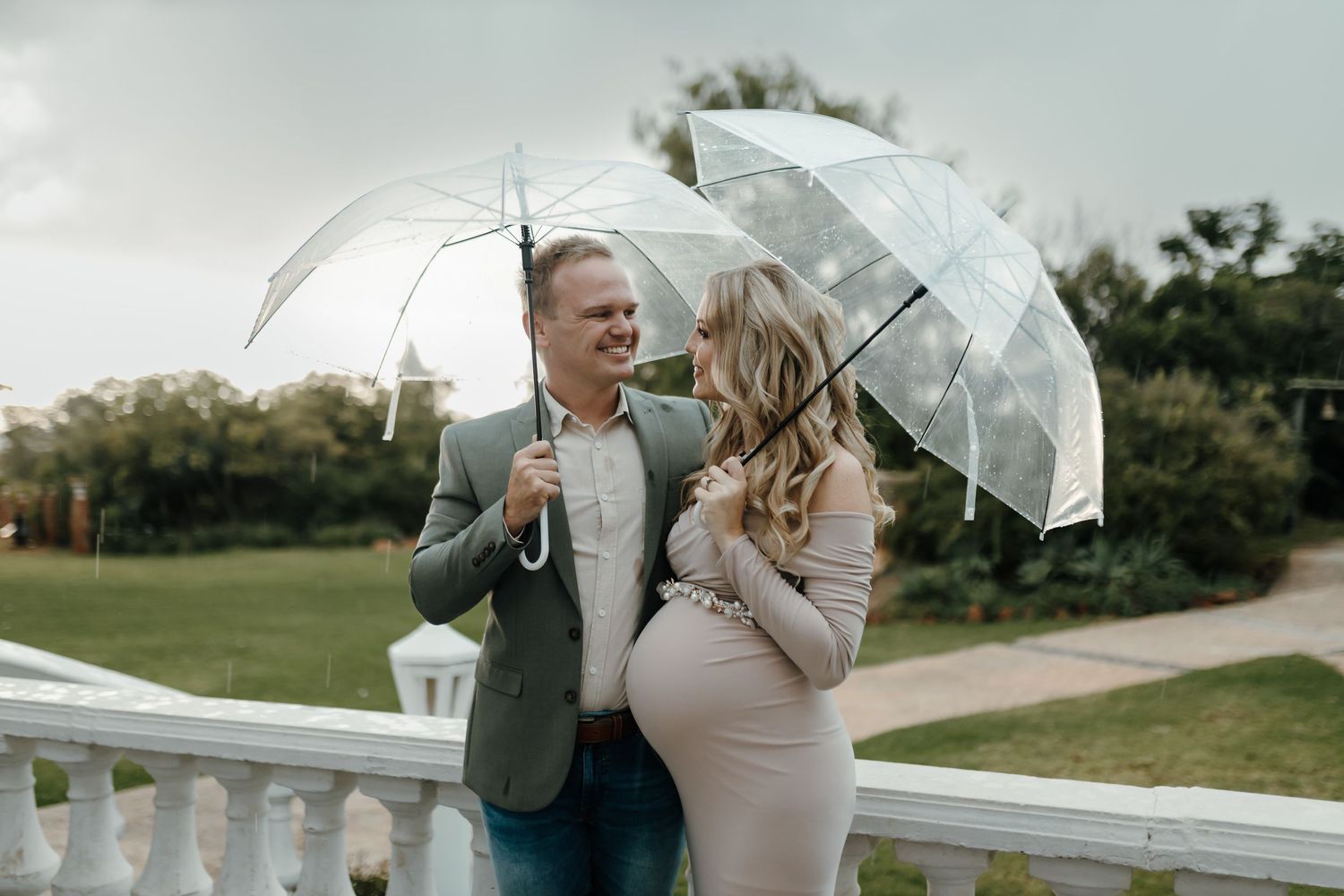 A couple with umbrellas share an intimate moment in front of white fencing on a rainy wedding day.