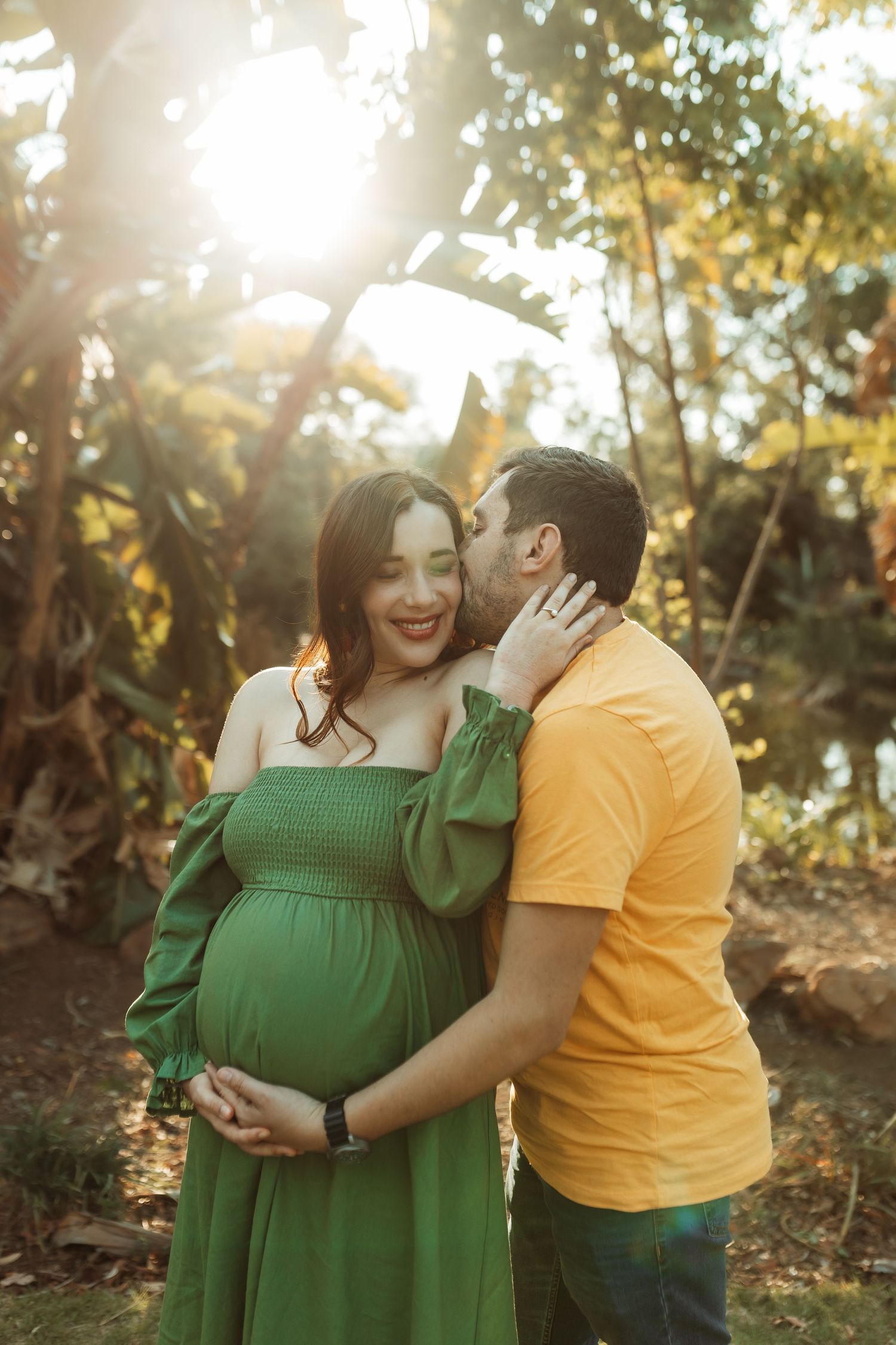 A loving couple in yellow and green outfits share tender moments in a sunlit forest setting during a pregnancy photoshoot.