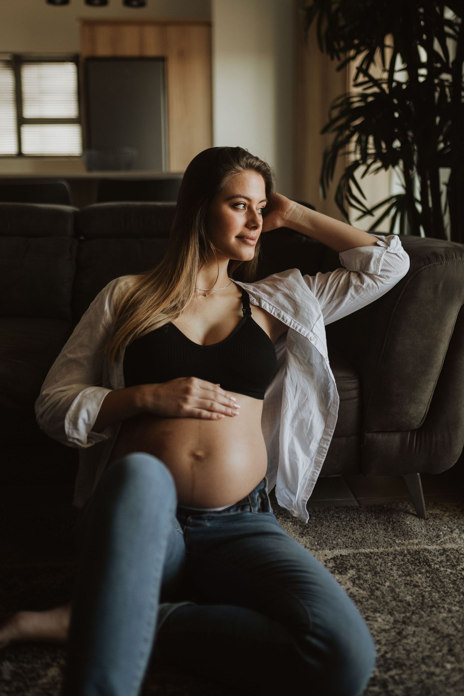 Expectant mother in white shirt shows baby bump while relaxing on gray couch in moody lighting.