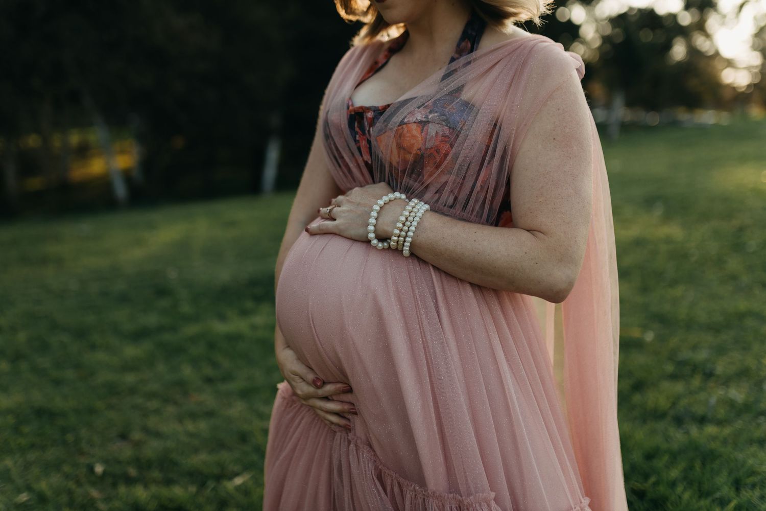 A person in a flowing blush pink dress cradles their baby bump against a sunlit grassy backdrop during sunset.