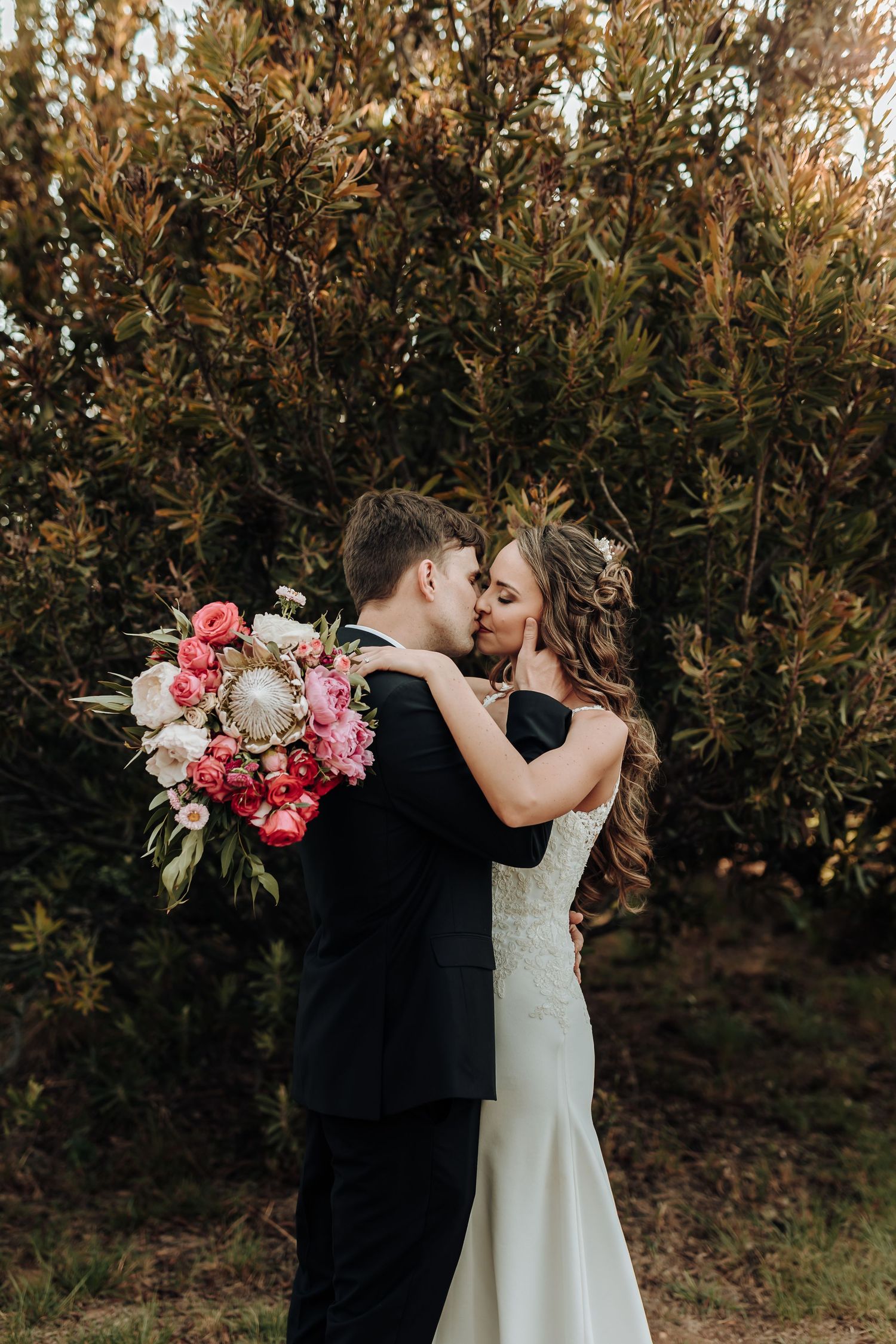 A bride and groom share a romantic kiss outdoors, surrounded by lush greenery, as she holds a vibrant pink and white bouquet.