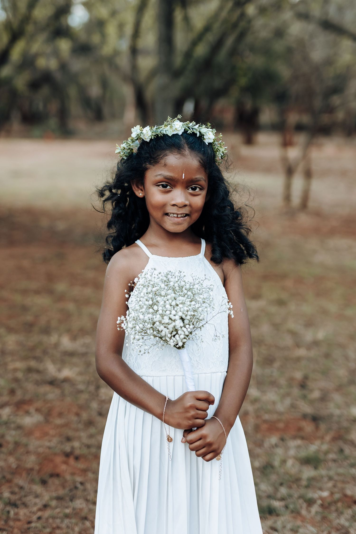 A young girl in a white dress wears a floral crown while standing in a wooded outdoor setting.