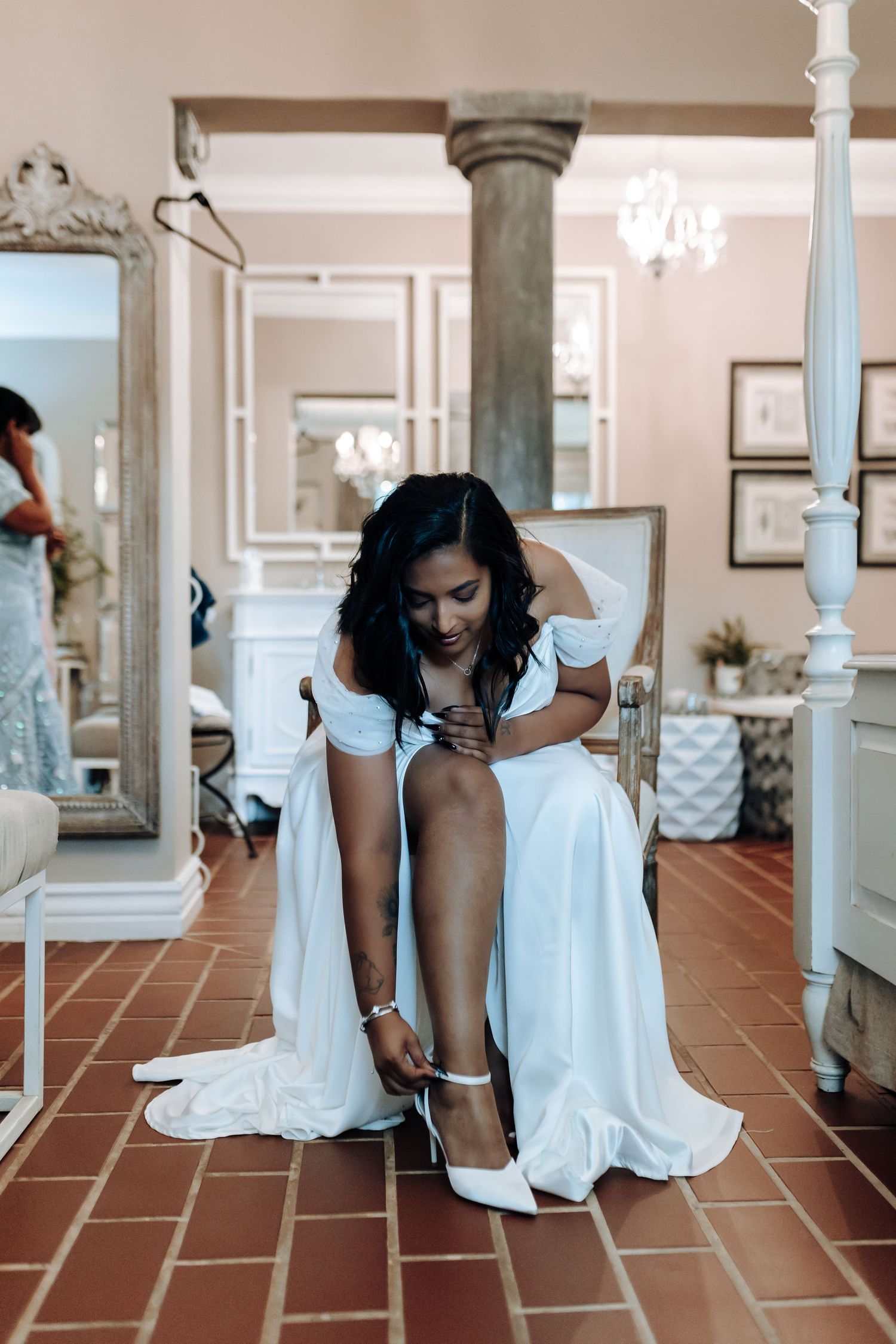 Bride in white dress putting on shoes while sitting on terracotta floor with elegant decor.