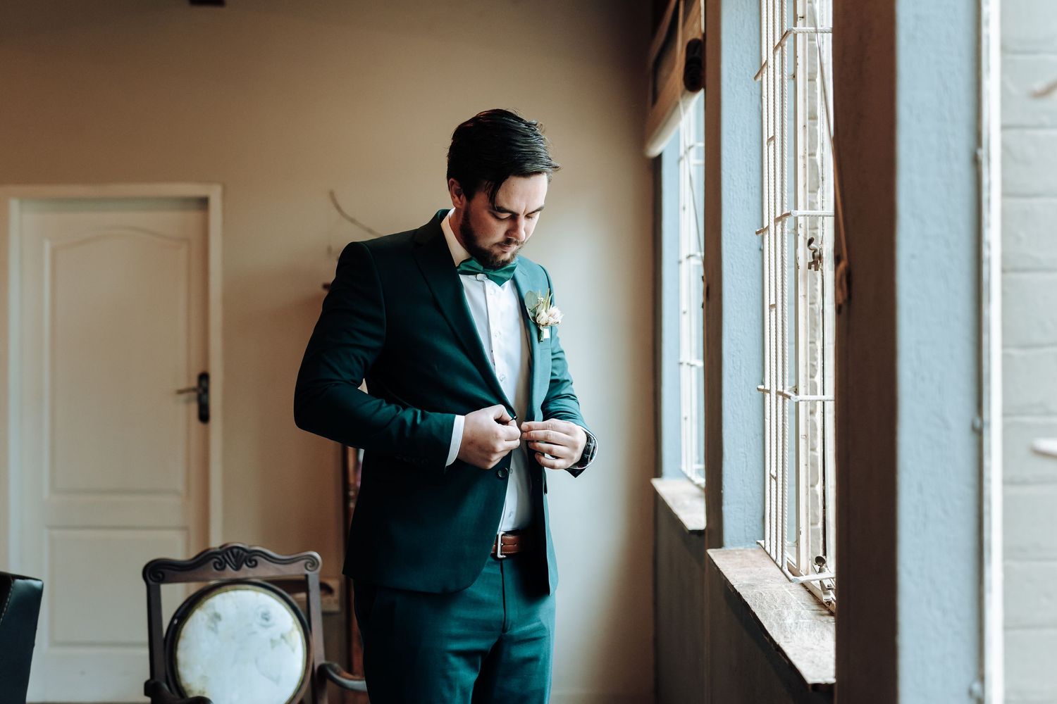 Emerald suit being adjusted by window with antique furniture visible in background.