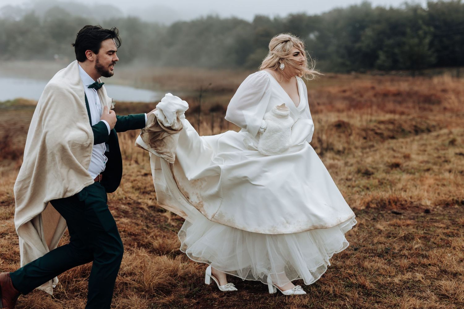 Wedding couple runs playfully through autumn field with flowing dress and cape caught in the wind.
