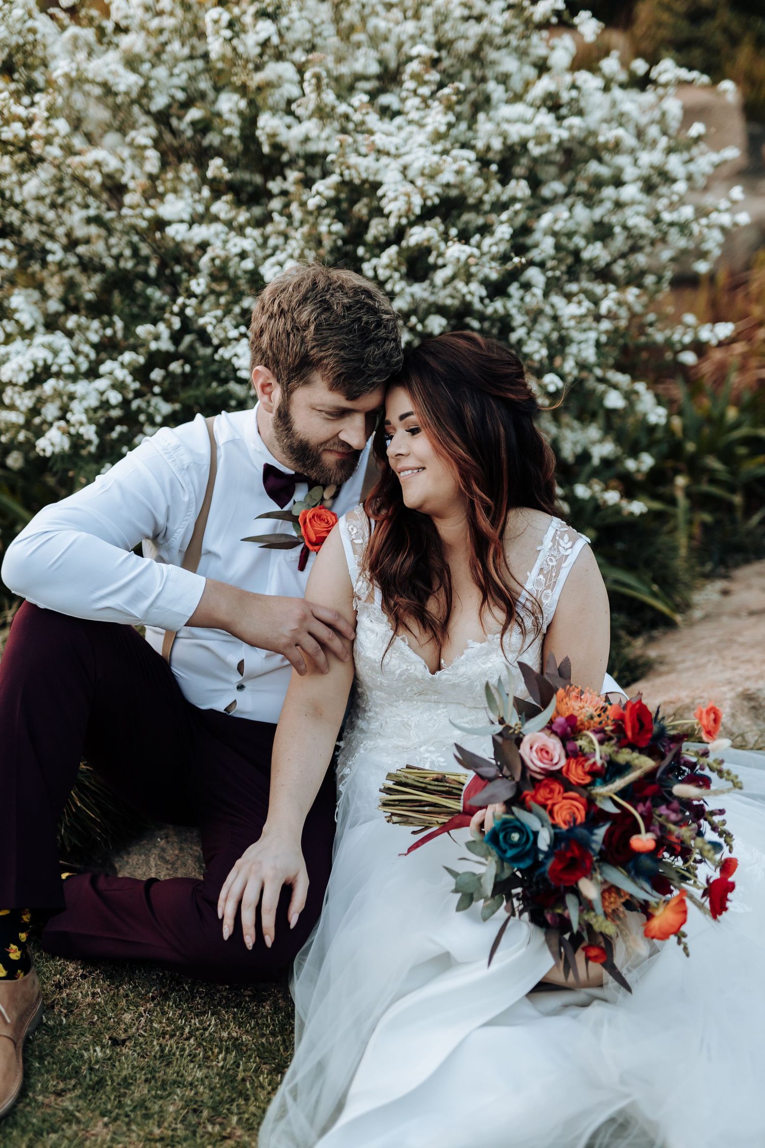 A romantic wedding scene with couple sitting by white flowering bushes and vibrant red bouquet.