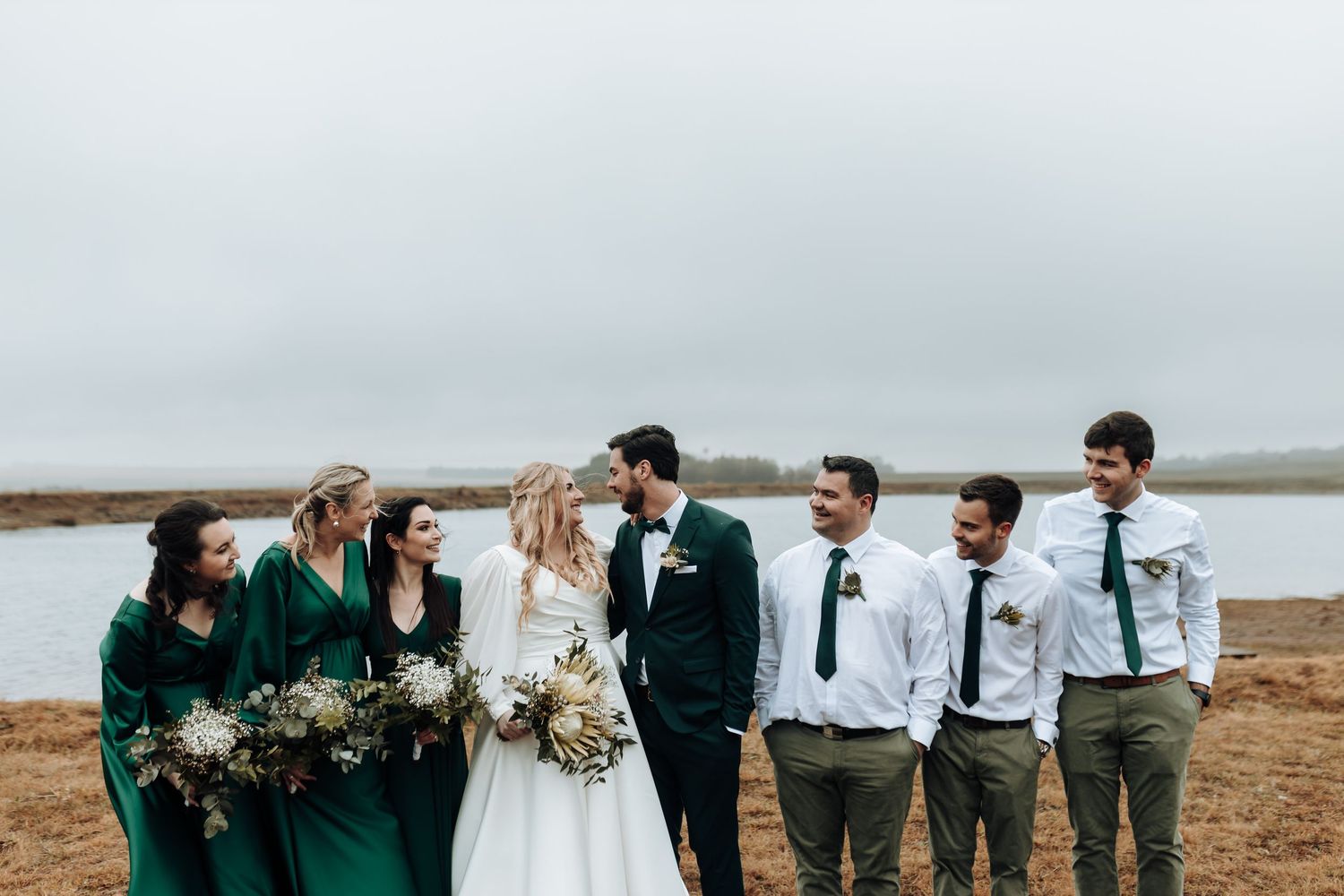 Wedding party in emerald green and white poses by waterfront on overcast day.