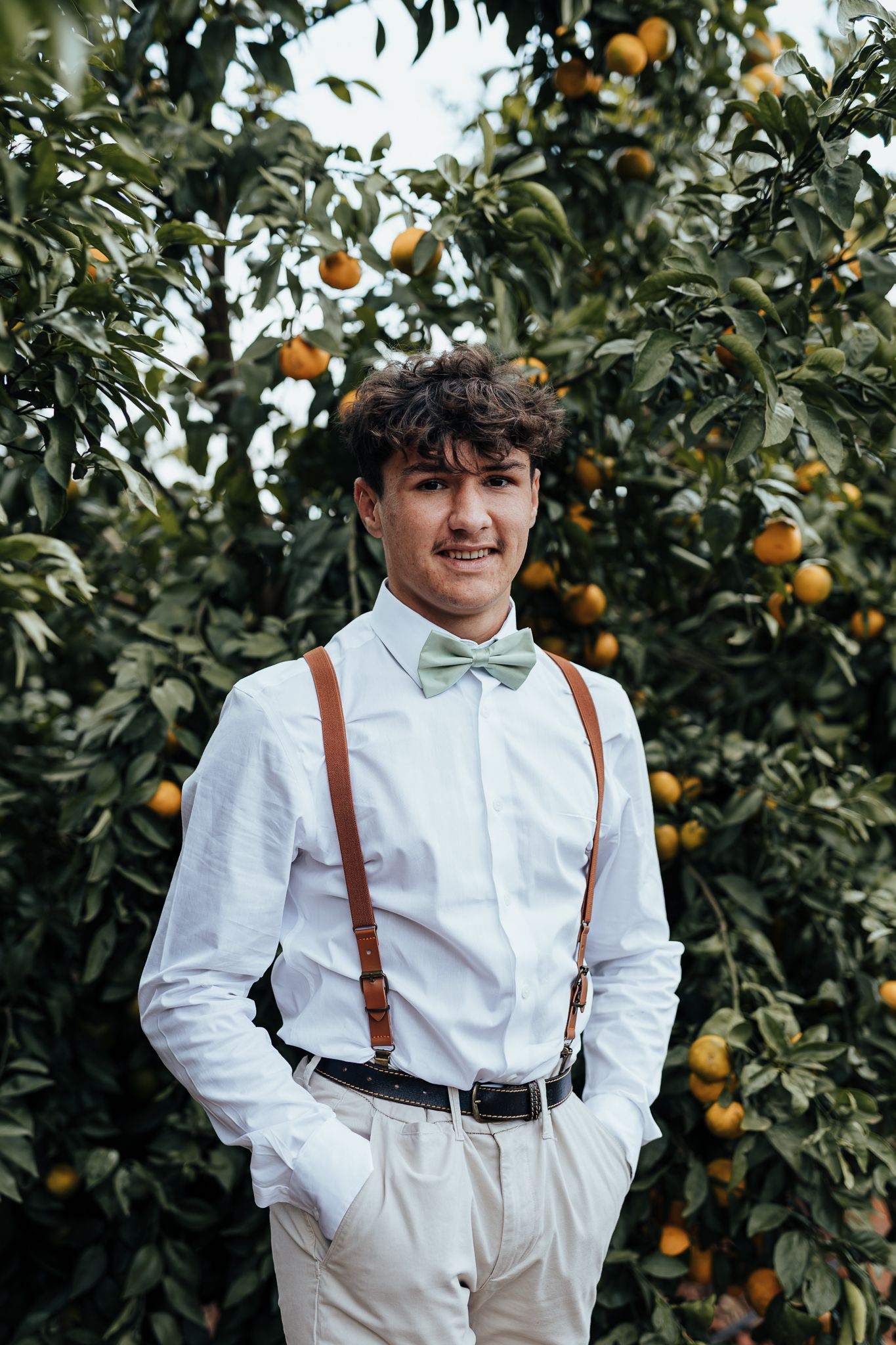 Wedding attendee in white shirt with bow tie and suspenders poses in citrus orchard.