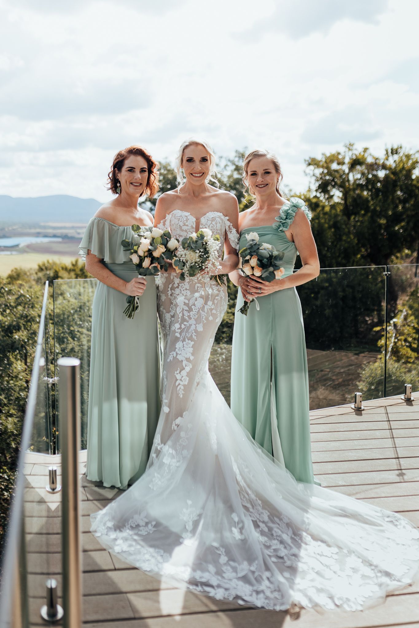 Bride in lace gown poses with two bridesmaids in sage green dresses on outdoor deck.
