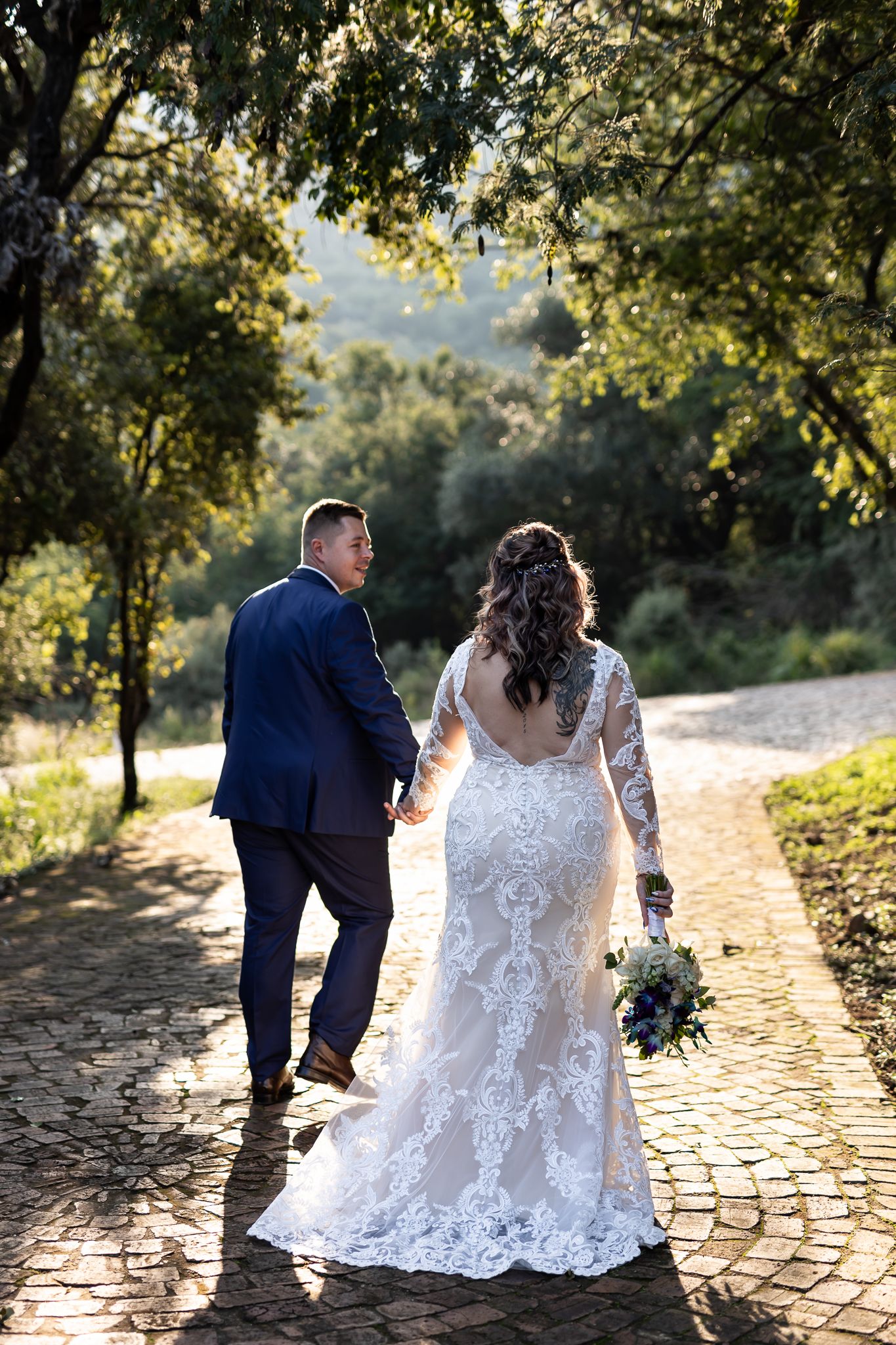 Newlyweds walk hand in hand on cobblestone path surrounded by lush greenery.