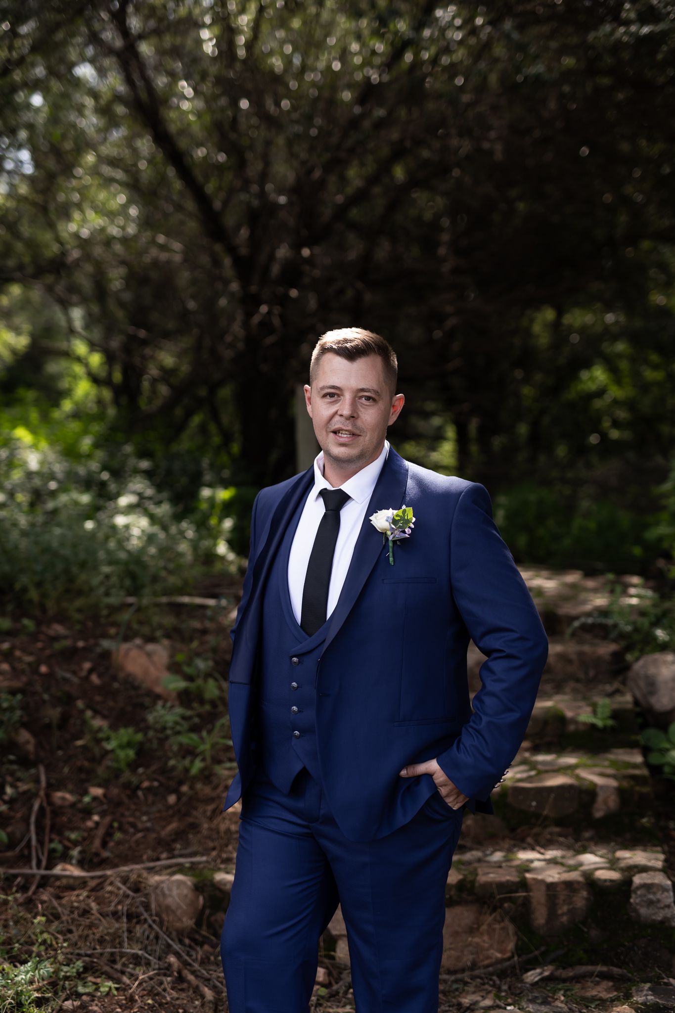 Groom in navy blue suit poses on stone steps with greenery background.