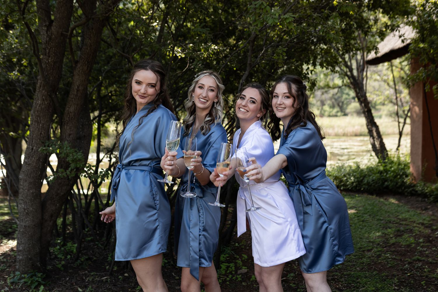 Wedding party in silk robes poses together under trees while holding drinks.