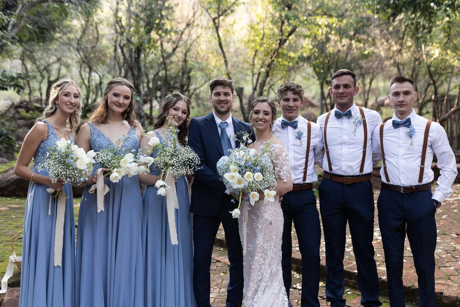 Wedding party poses outdoors in light blue bridesmaid dresses and navy suspenders against natural backdrop.