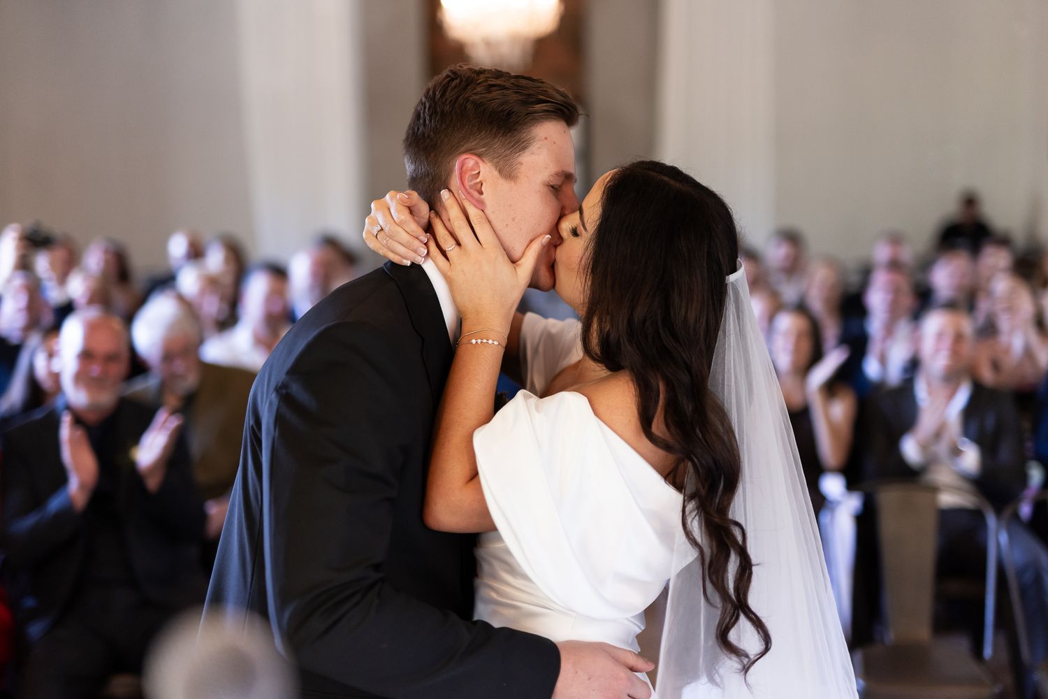 A couple sharing their first kiss as newlyweds surrounded by guests at their wedding ceremony.