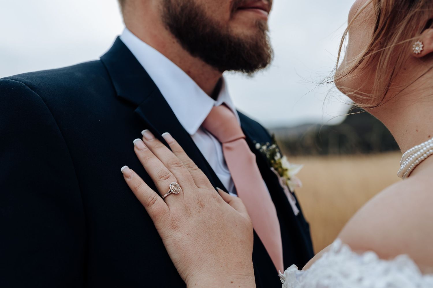Close up wedding photo showing hands wearing rings and formal attire during an outdoor ceremony.