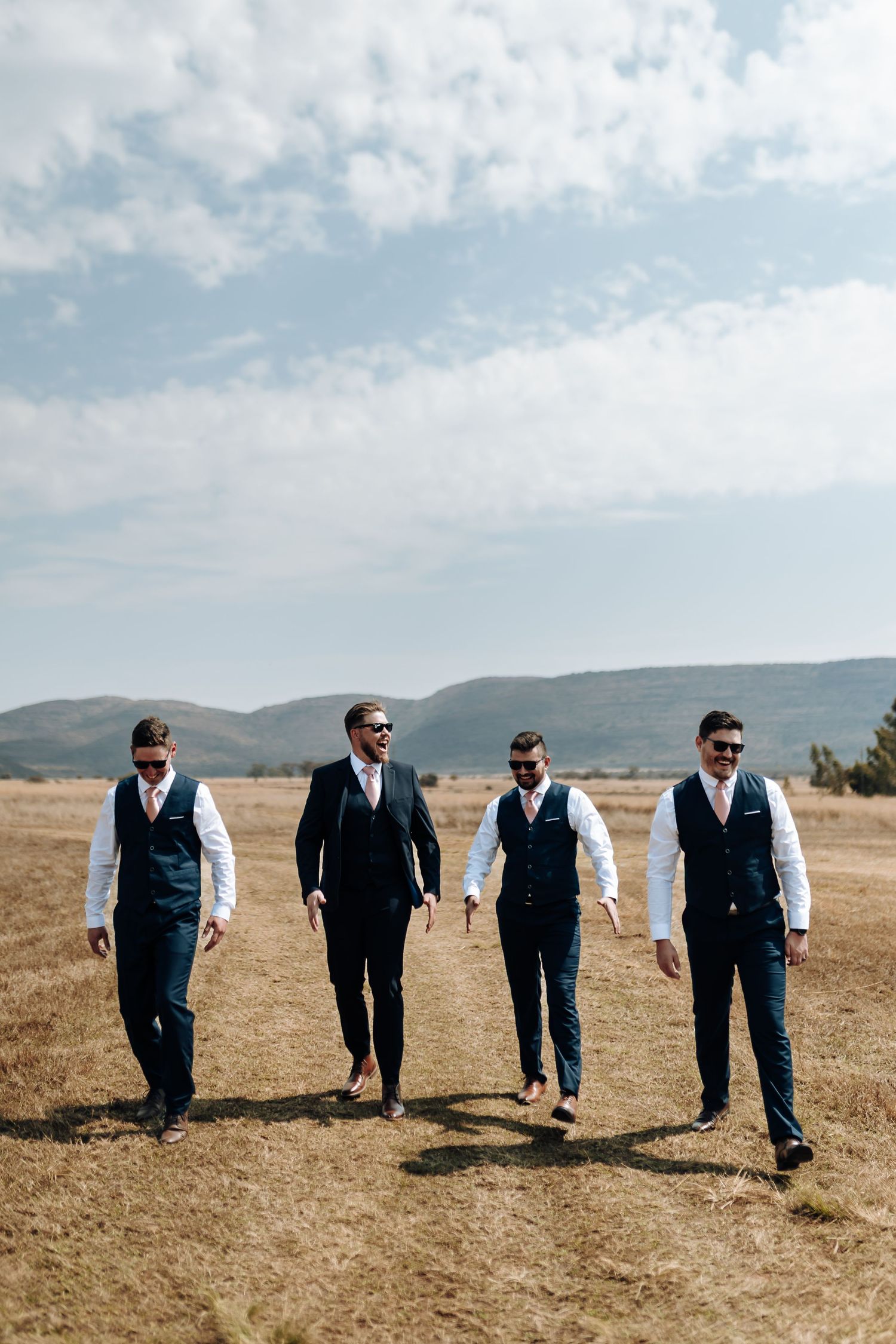 A group in formal attire with matching vests walk together across a mountainous landscape under partly cloudy skies.