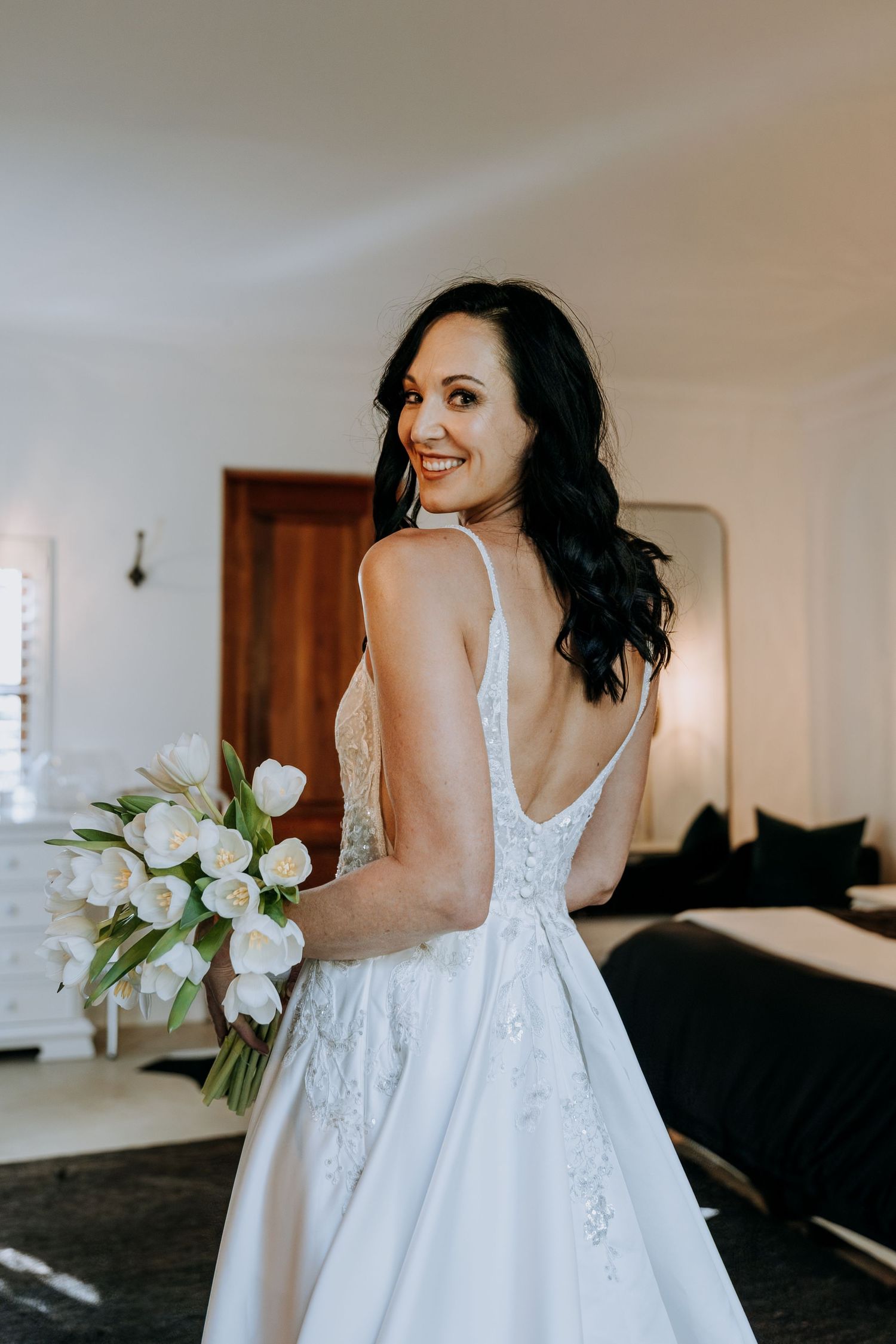 Smiling bride in a white backless lace wedding gown holding a white tulip bouquet, posing indoors.