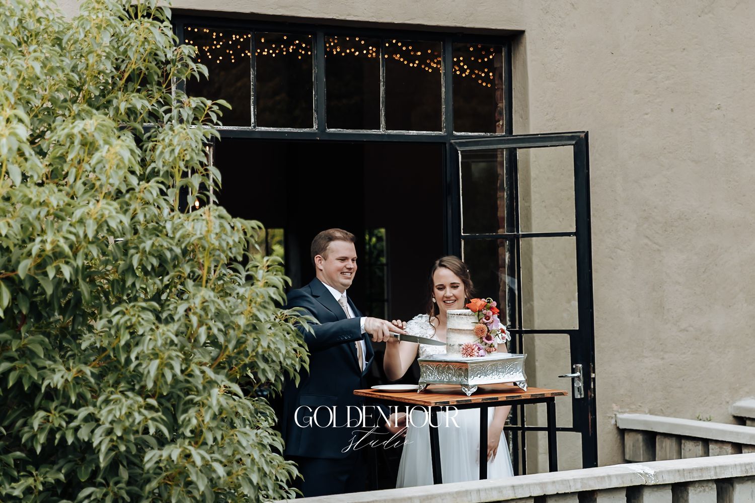 Smiling bride and groom cut their wedding cake together on an outdoor balcony.