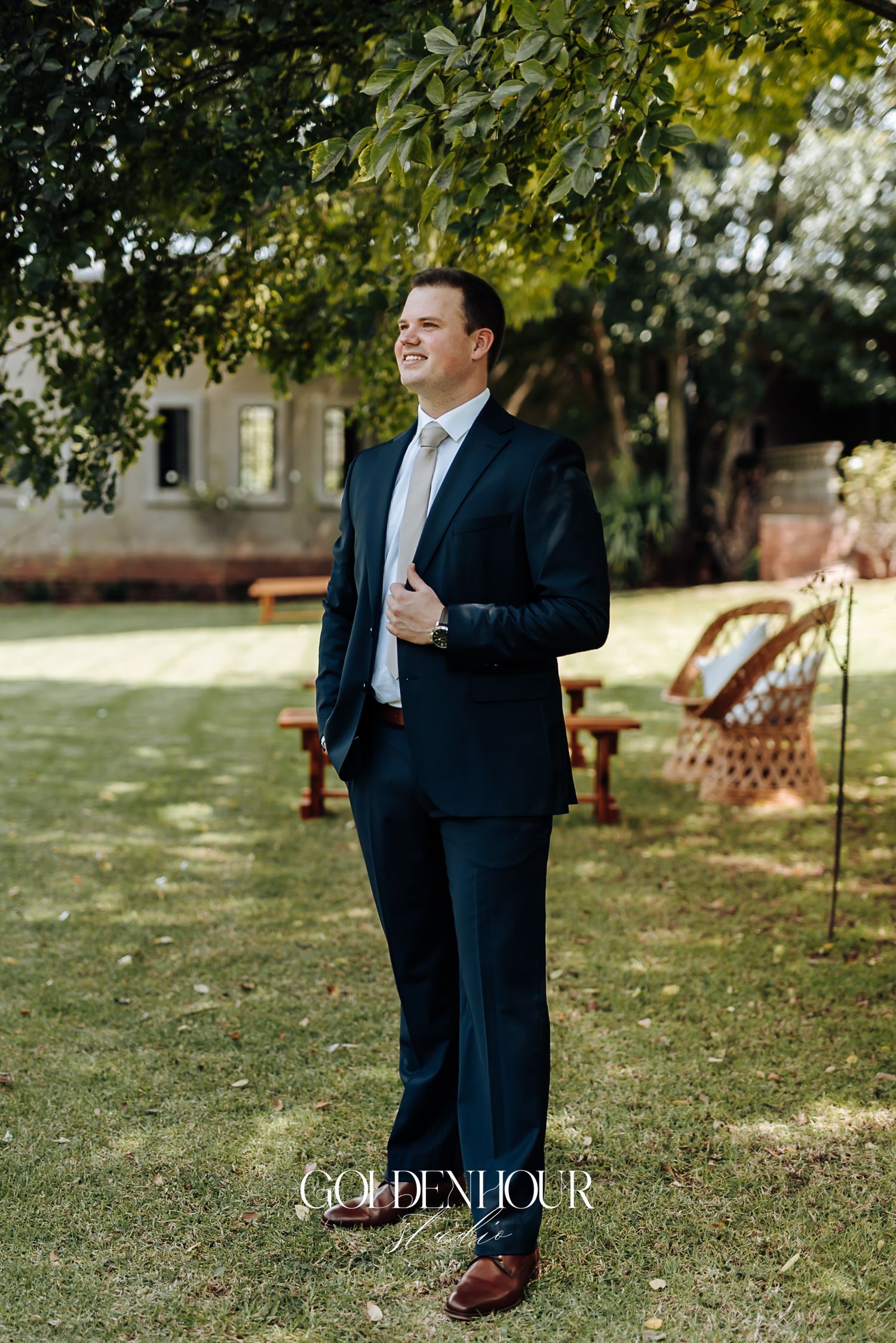 Groom in a navy suit smiling outdoors under a tree at a garden wedding venue.