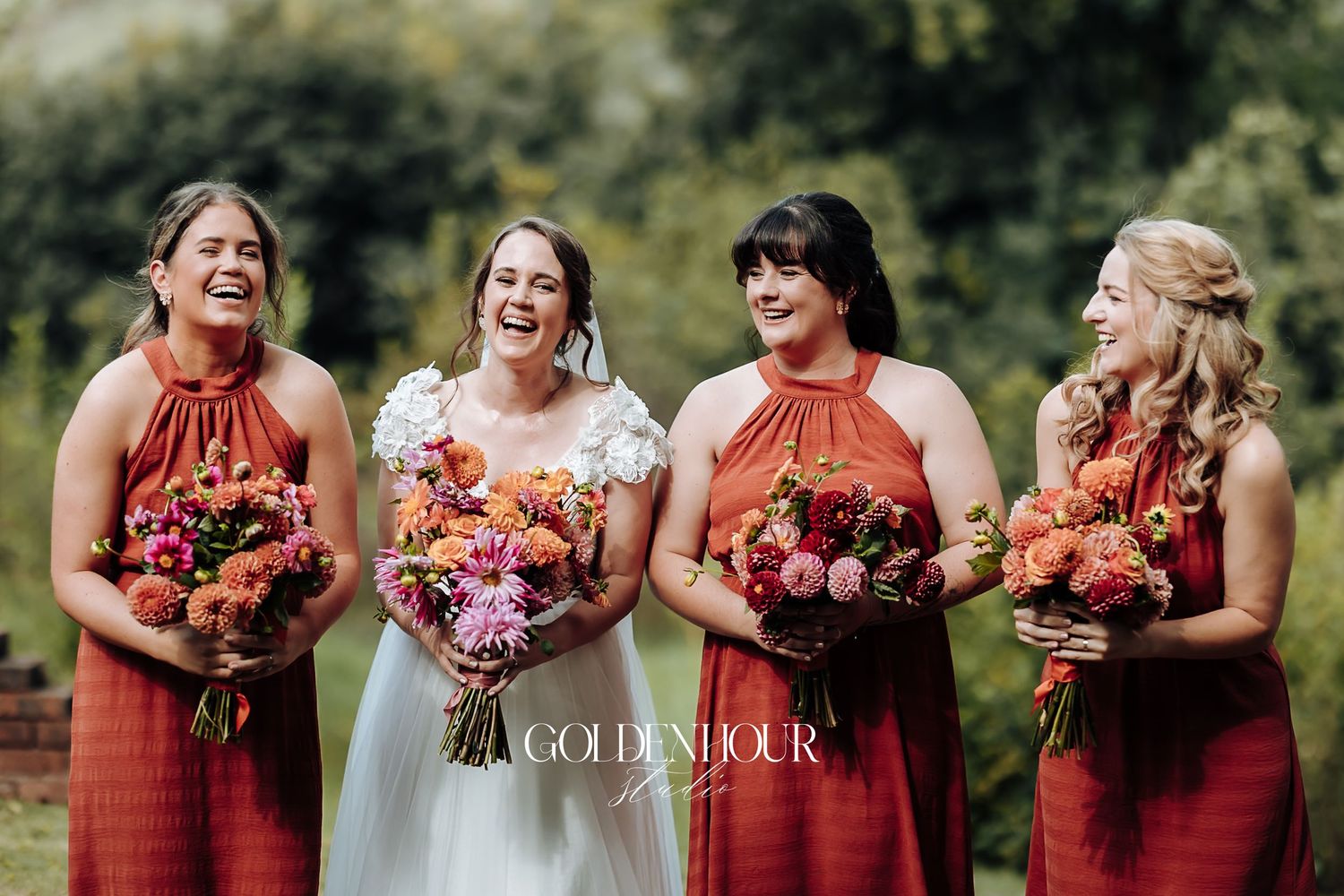 Bride and bridesmaids in rust-colored dresses laughing together outdoors holding vibrant pink and orange bouquets.