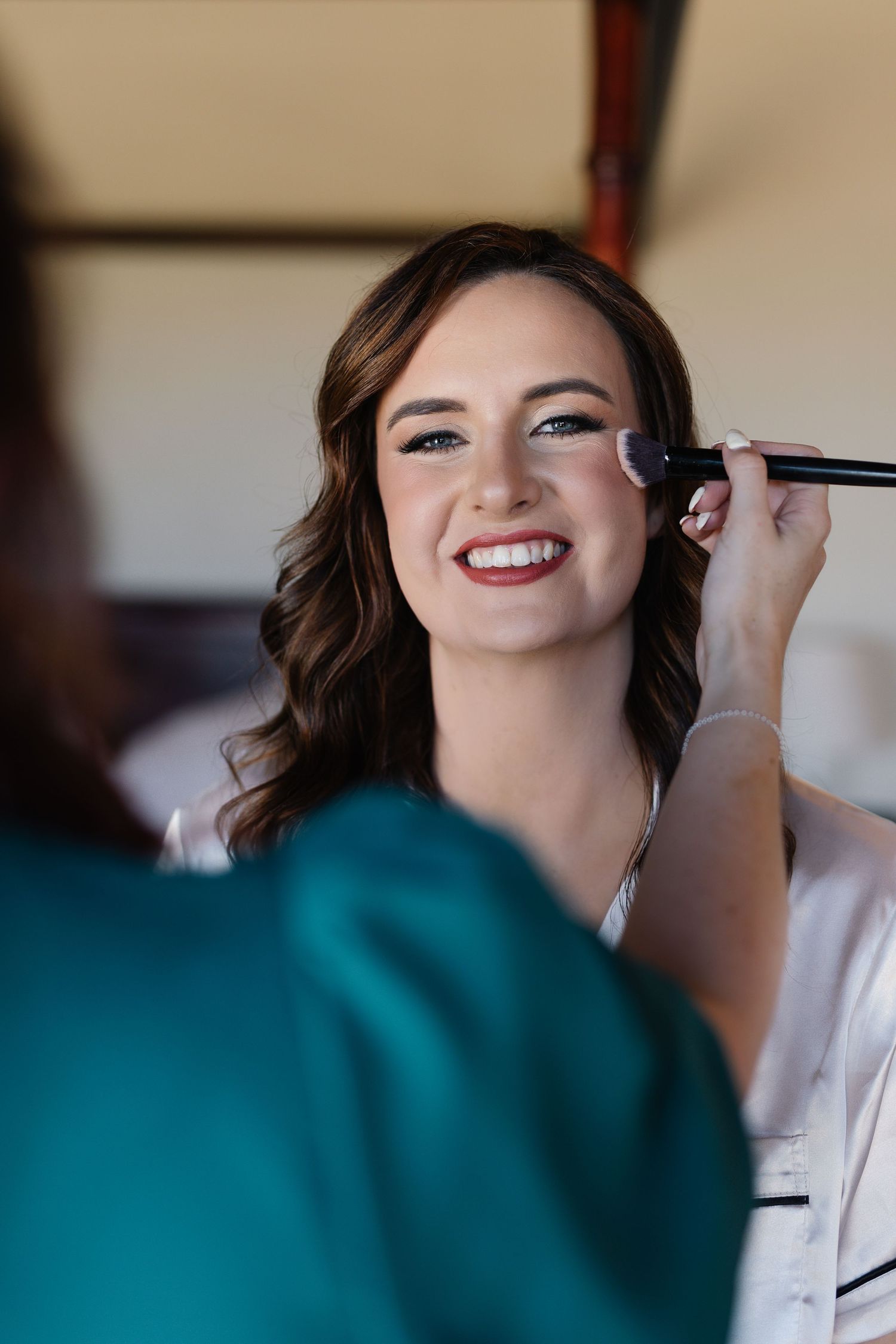 Makeup artist applies eyeshadow to smiling client wearing white robe during wedding preparation session.