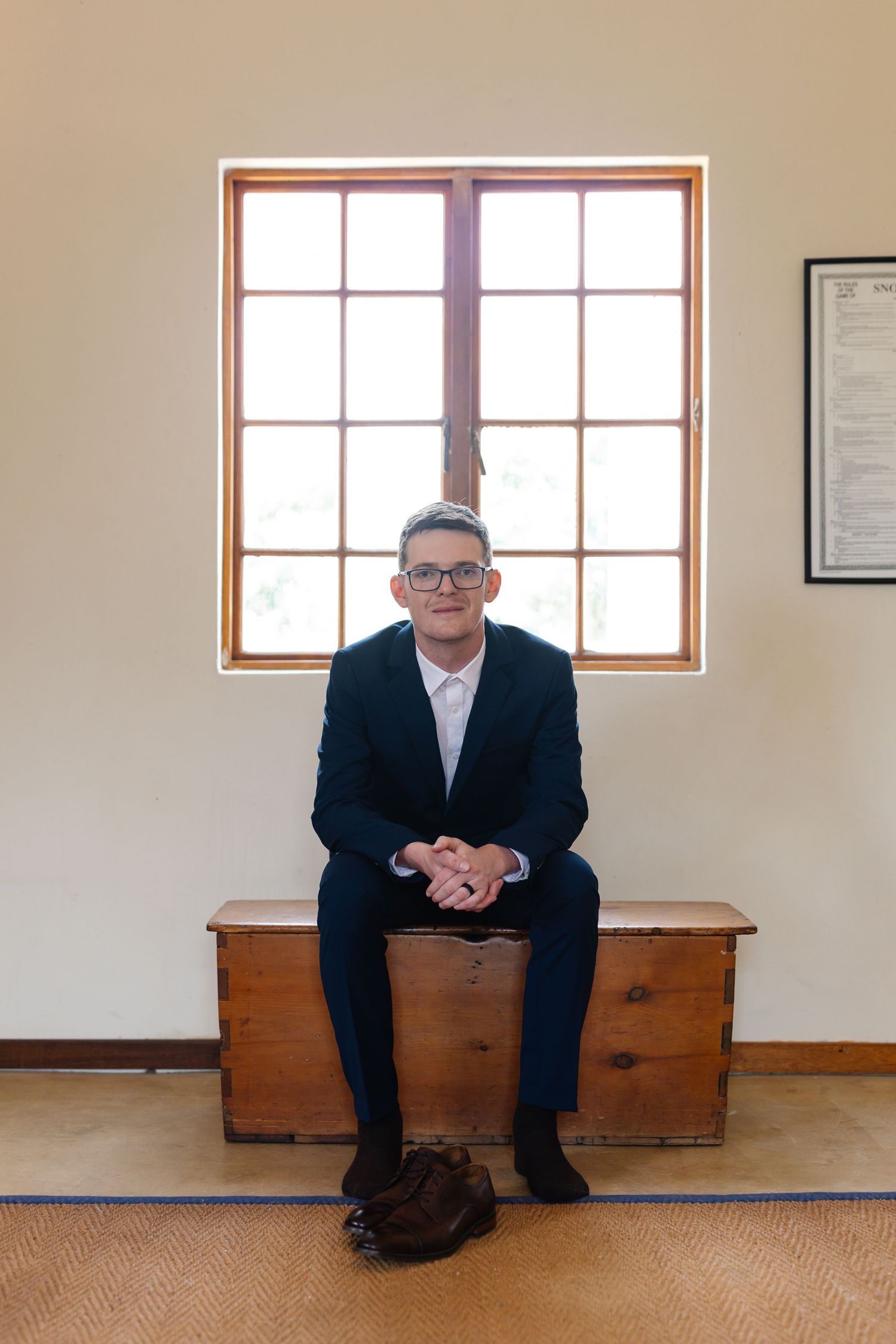 A person in a dark suit sits on a wooden bench in a minimalist room with windows and beige walls.