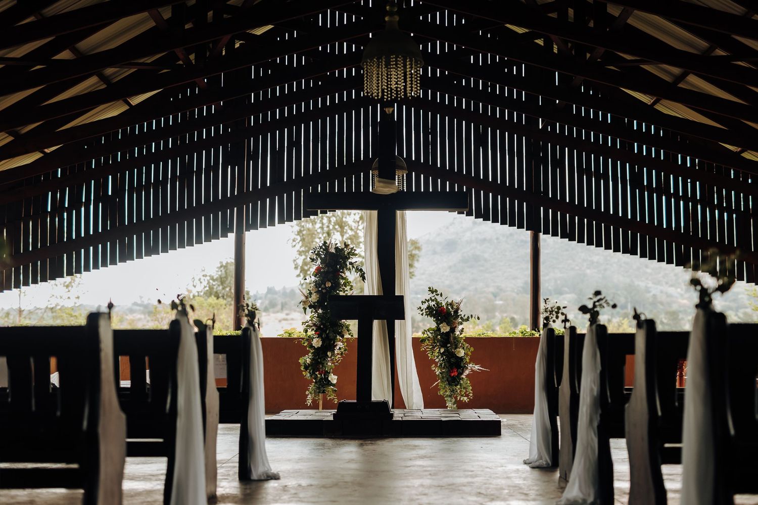 Interior view of a rustic wooden barn or pavilion with exposed beams and hanging floral arrangements.