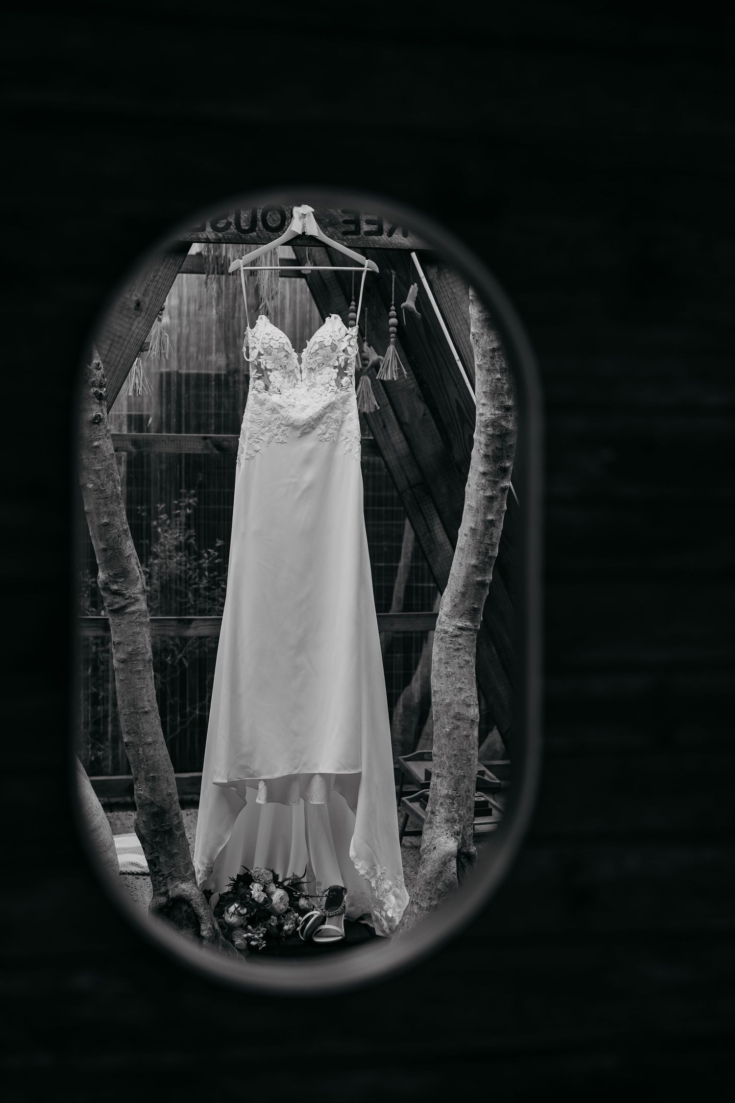 A wedding dress hangs elegantly between trees in a moody black and white photograph with circular vignette framing.