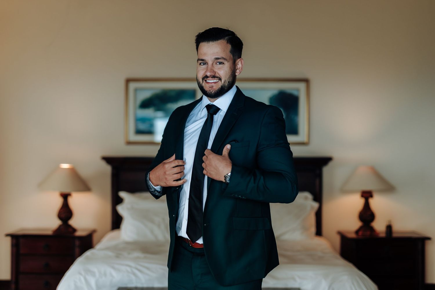 Professional in a dark suit and tie adjusts his jacket while standing in a warmly lit hotel room.