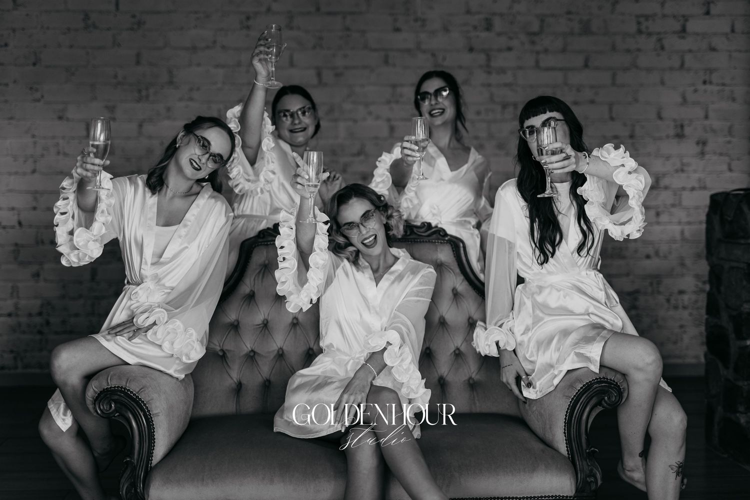Wedding party members in white robes laugh and celebrate together while sitting on a vintage couch.