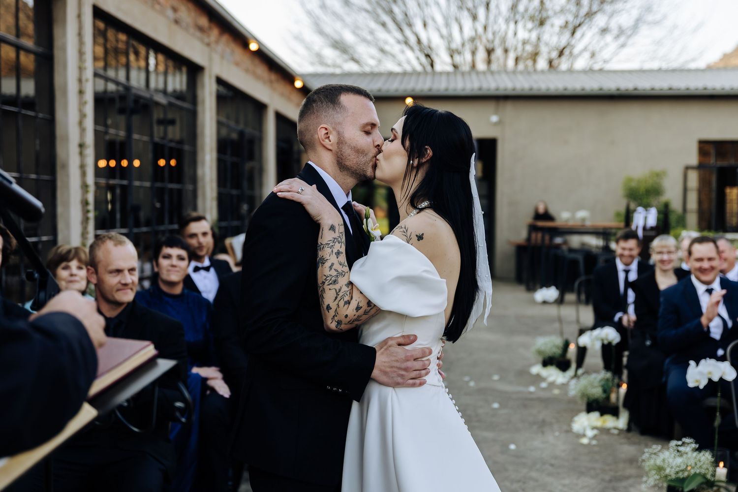 A couple embraces during their outdoor wedding ceremony while guests look on at a rustic venue.