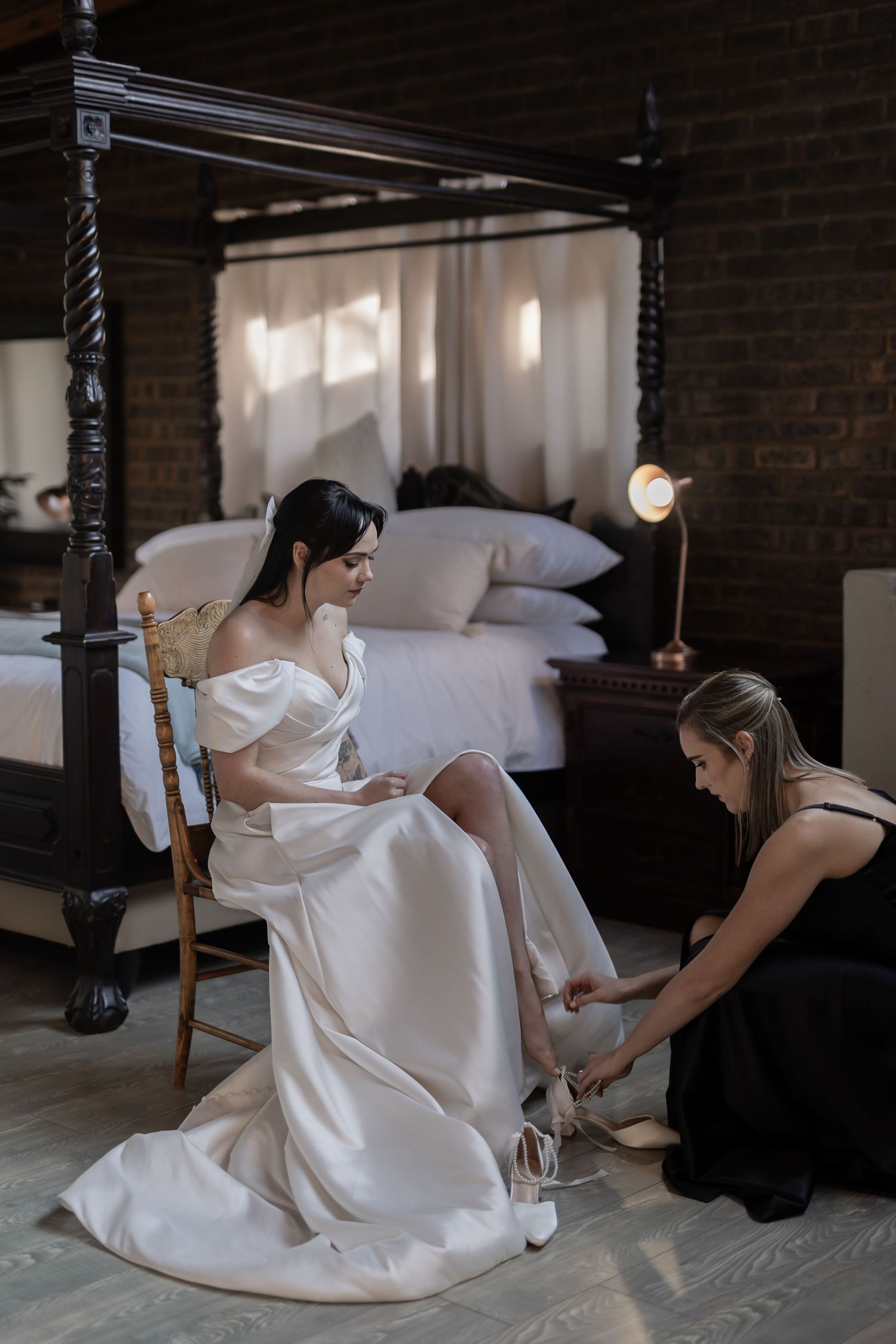 A woman in an elegant white wedding dress sits in a cushioned chair while someone helps with her shoes in a rustic bedroom.
