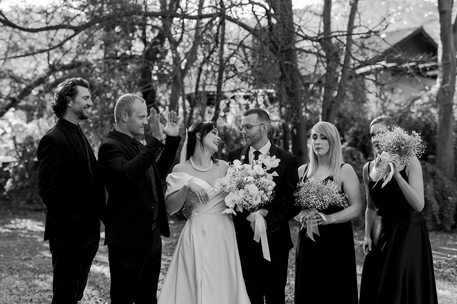 A black and white photo of a wedding party outdoors with bridesmaids in black dresses and white floral bouquets.