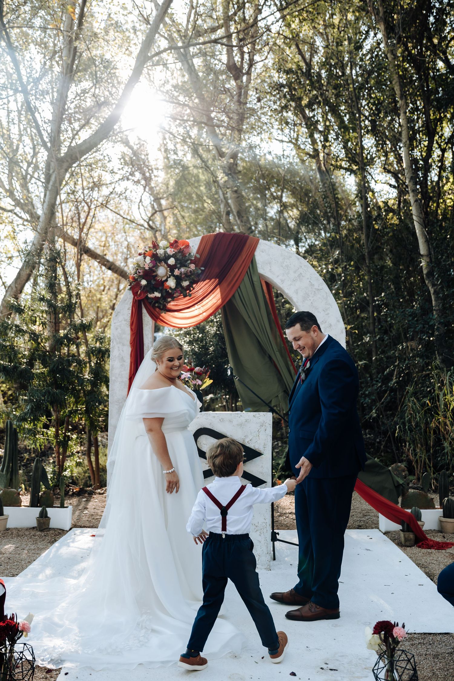 Outdoor wedding ceremony with bride in white gown, groom in navy suit, and young boy holding hands under floral arch.