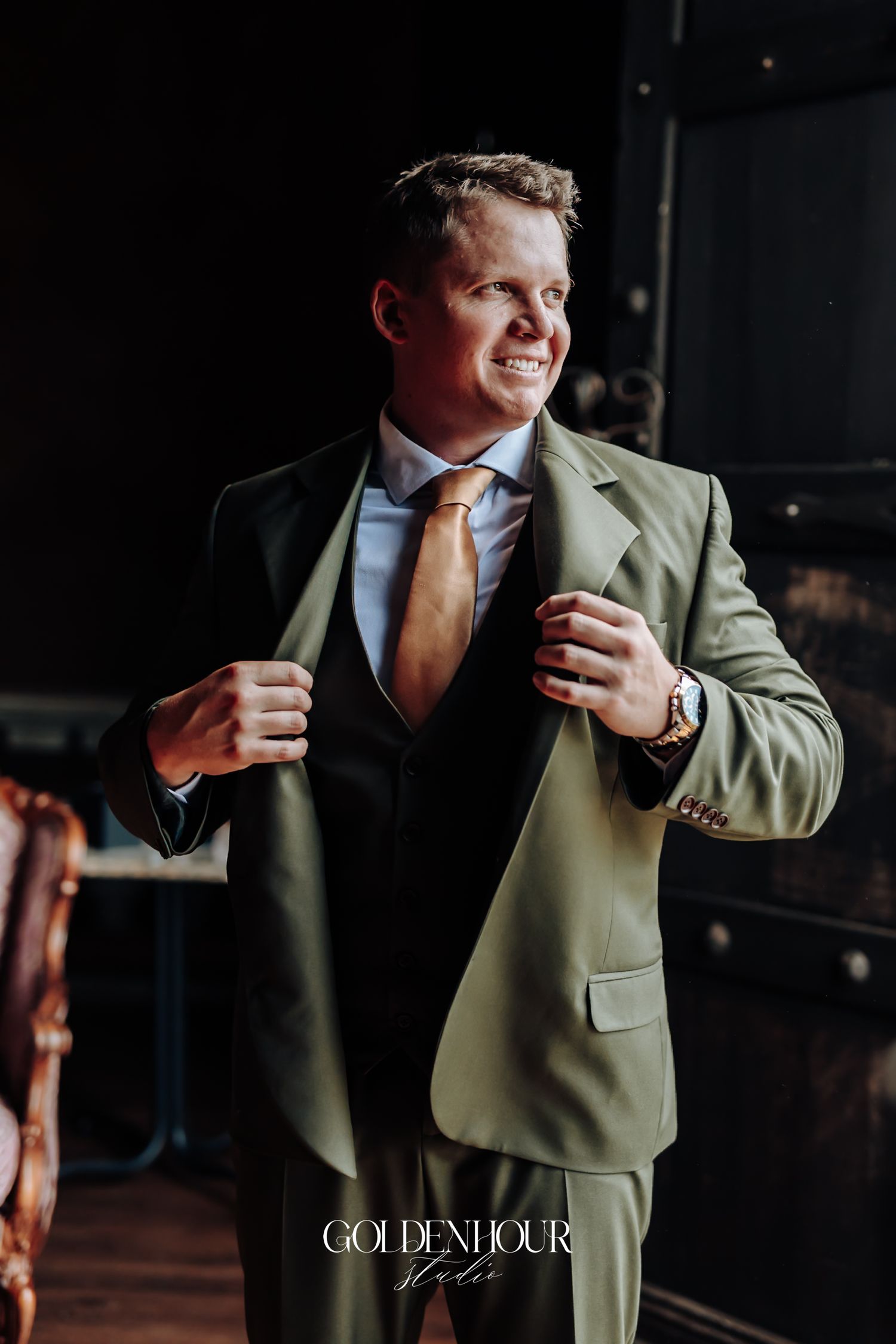 Smiling groom in olive green suit adjusting jacket lapels in dark elegant interior.