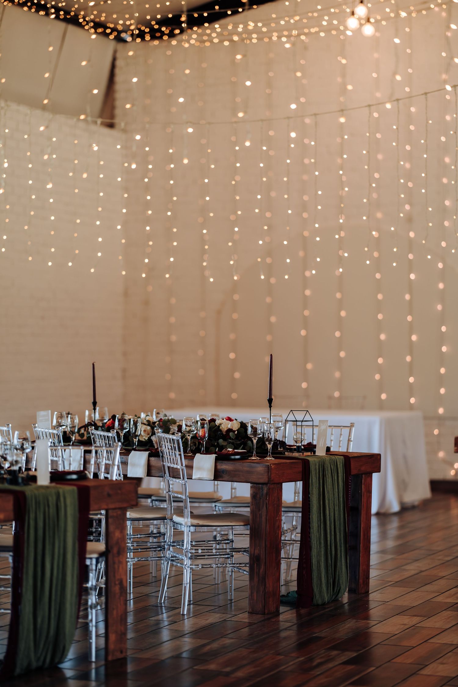 Elegant wedding reception with fairy light backdrop, wooden tables, green draping, and ghost chairs on hardwood floors.
